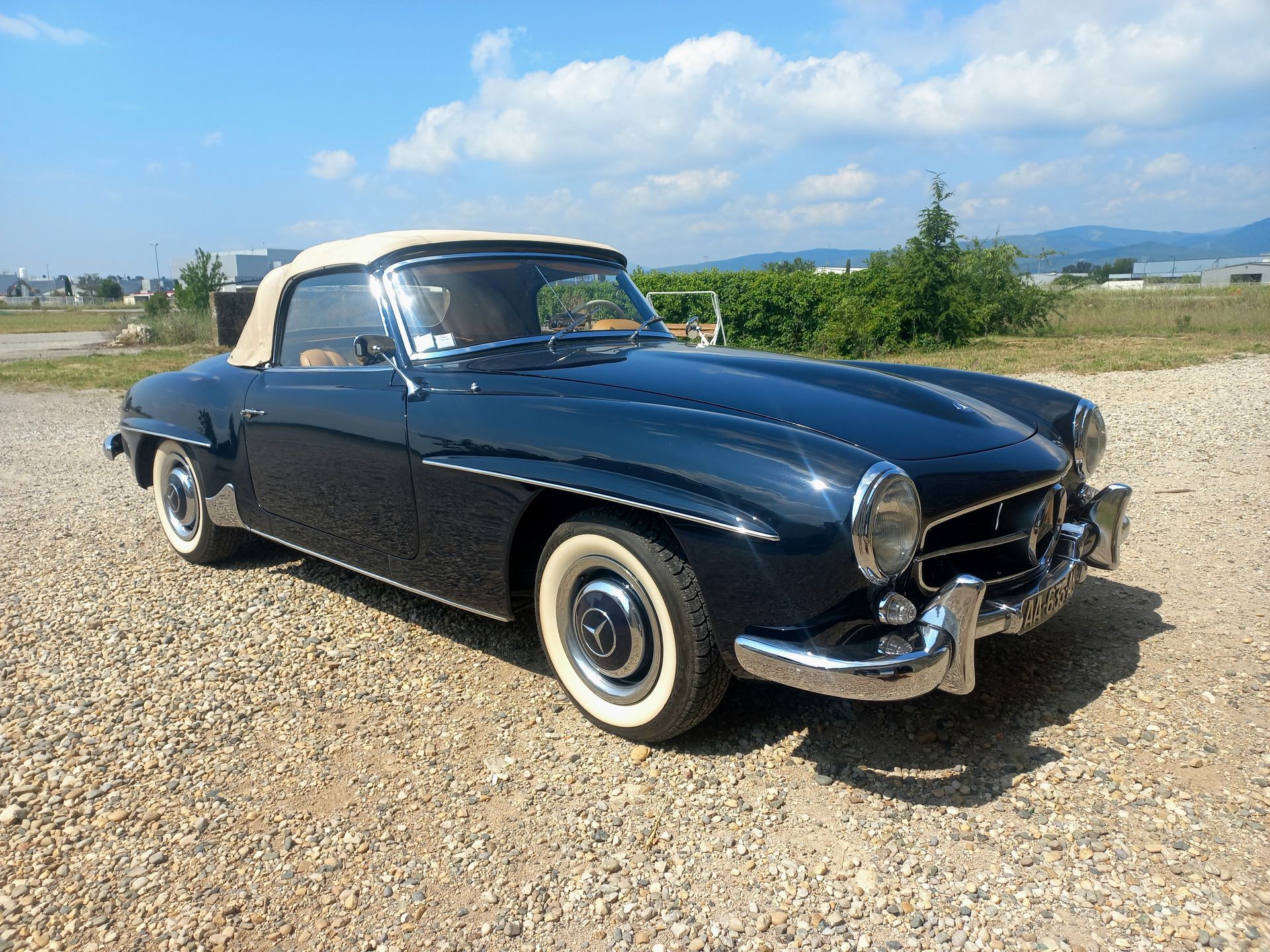 Une voiture décapotable noire vintage garée sur du gravier sous un ciel lumineux, avec des montagnes à l'horizon.