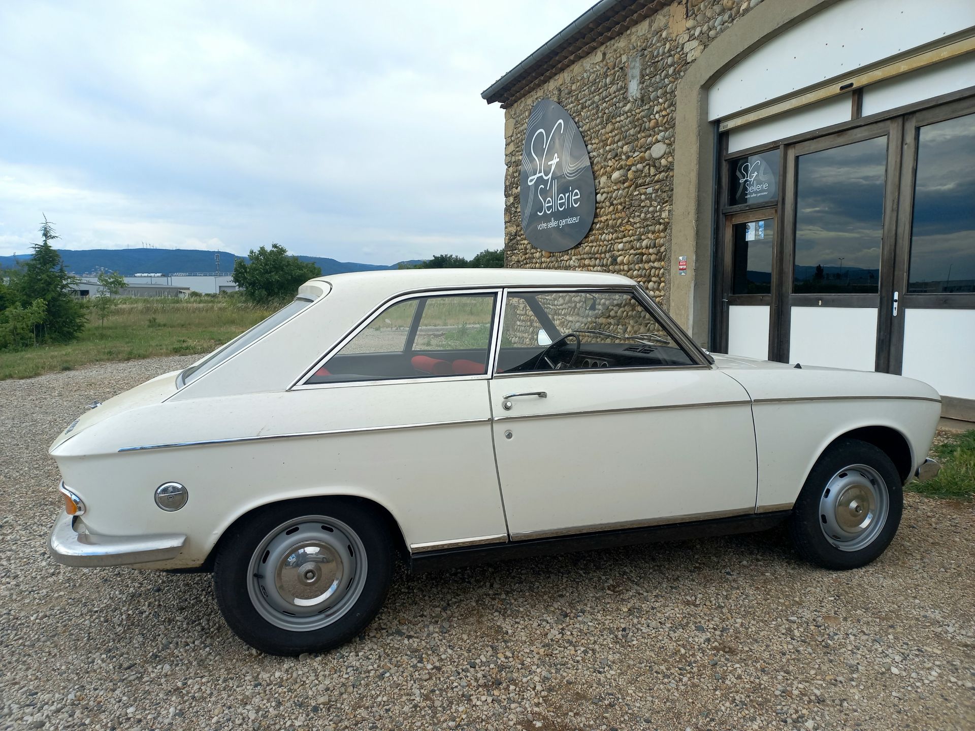 Un coupé blanc ancien est garé à côté d'un bâtiment en pierre avec une porte de garage, sur un terrain gravillonné.