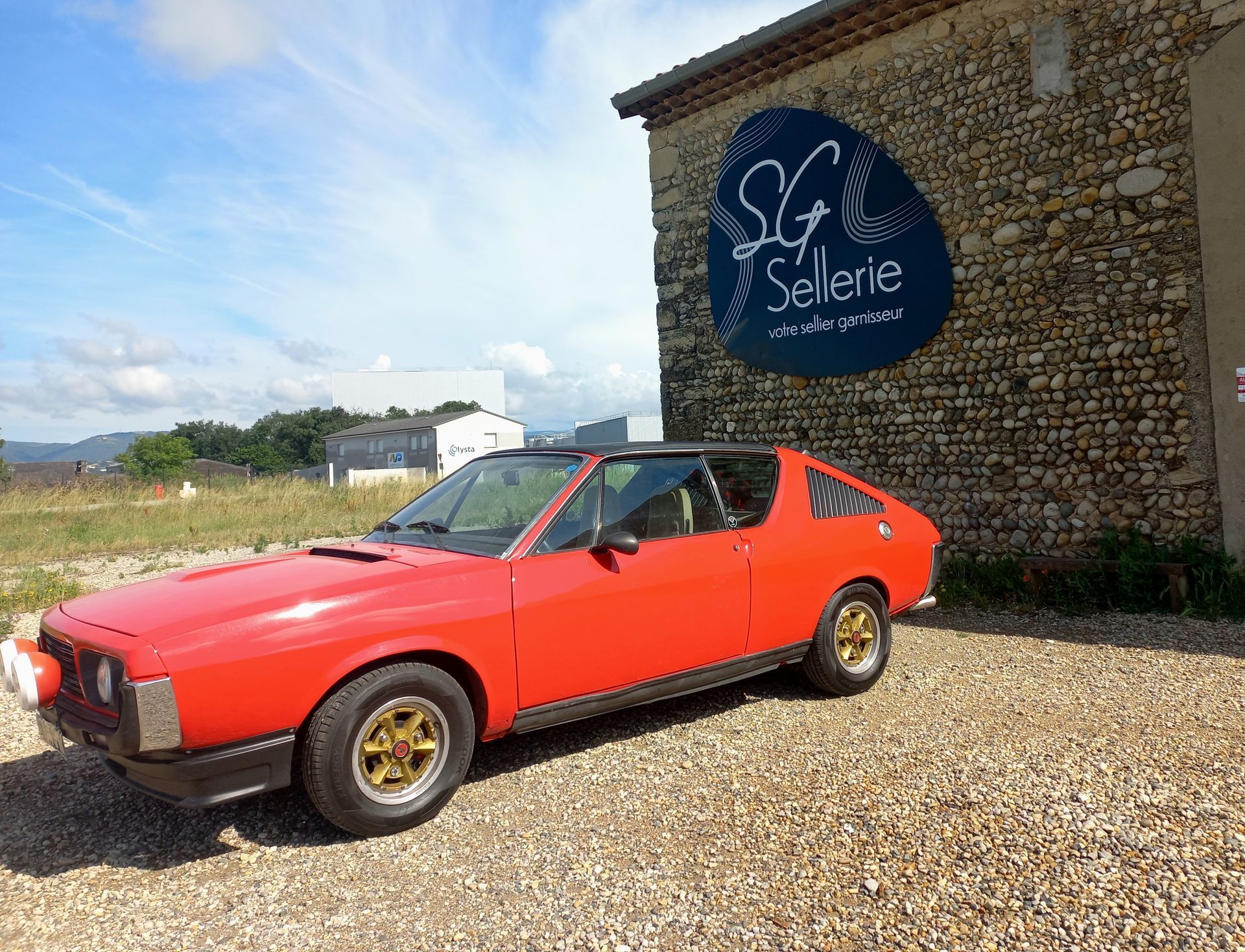 Voiture de sport classique rouge garée à côté d'un bâtiment en pierre avec un panneau noir sur un terrain gravillonné.