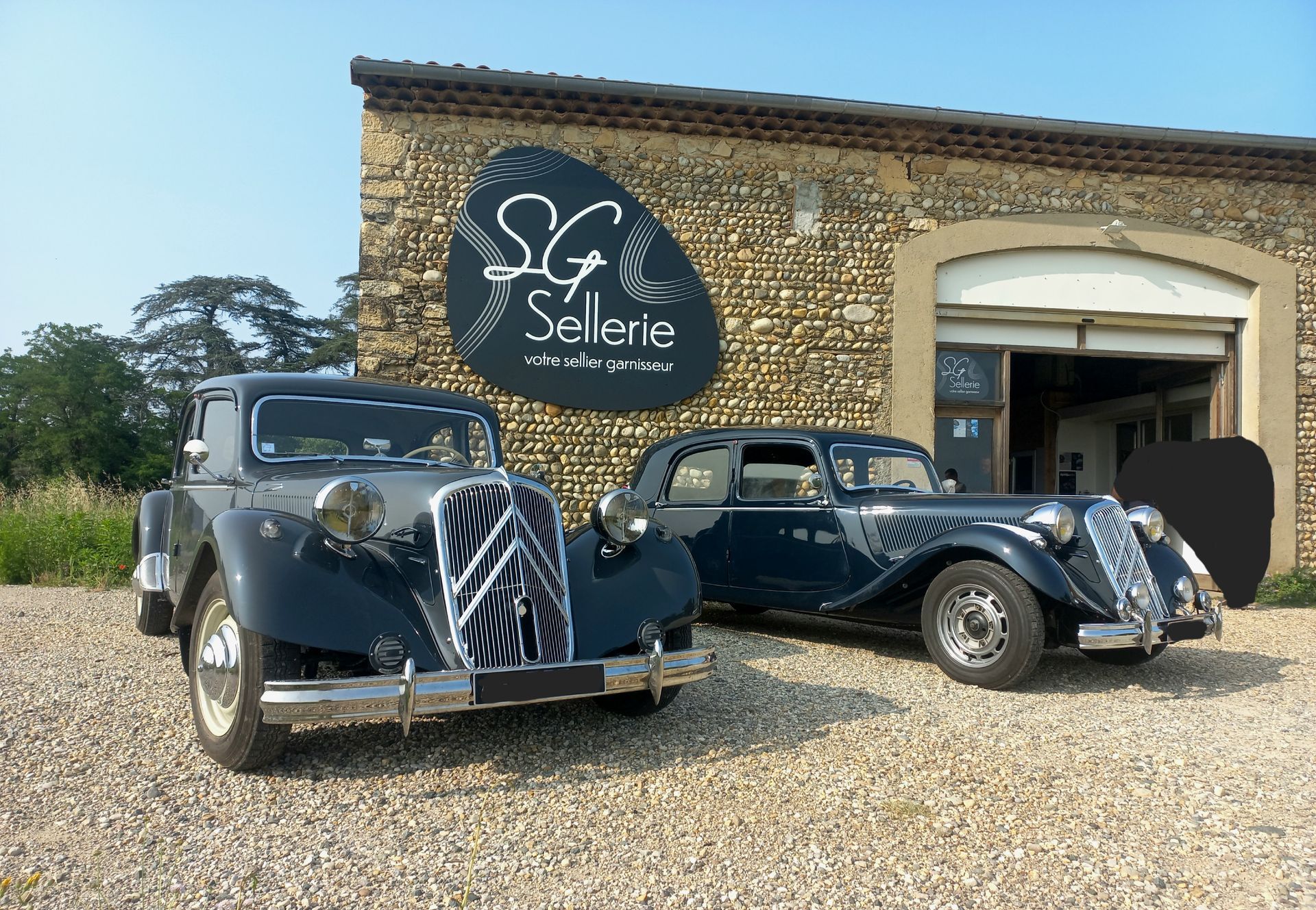 Deux voitures noires classiques garées devant un bâtiment en pierre orné d'une grande enseigne SG Sellerie.