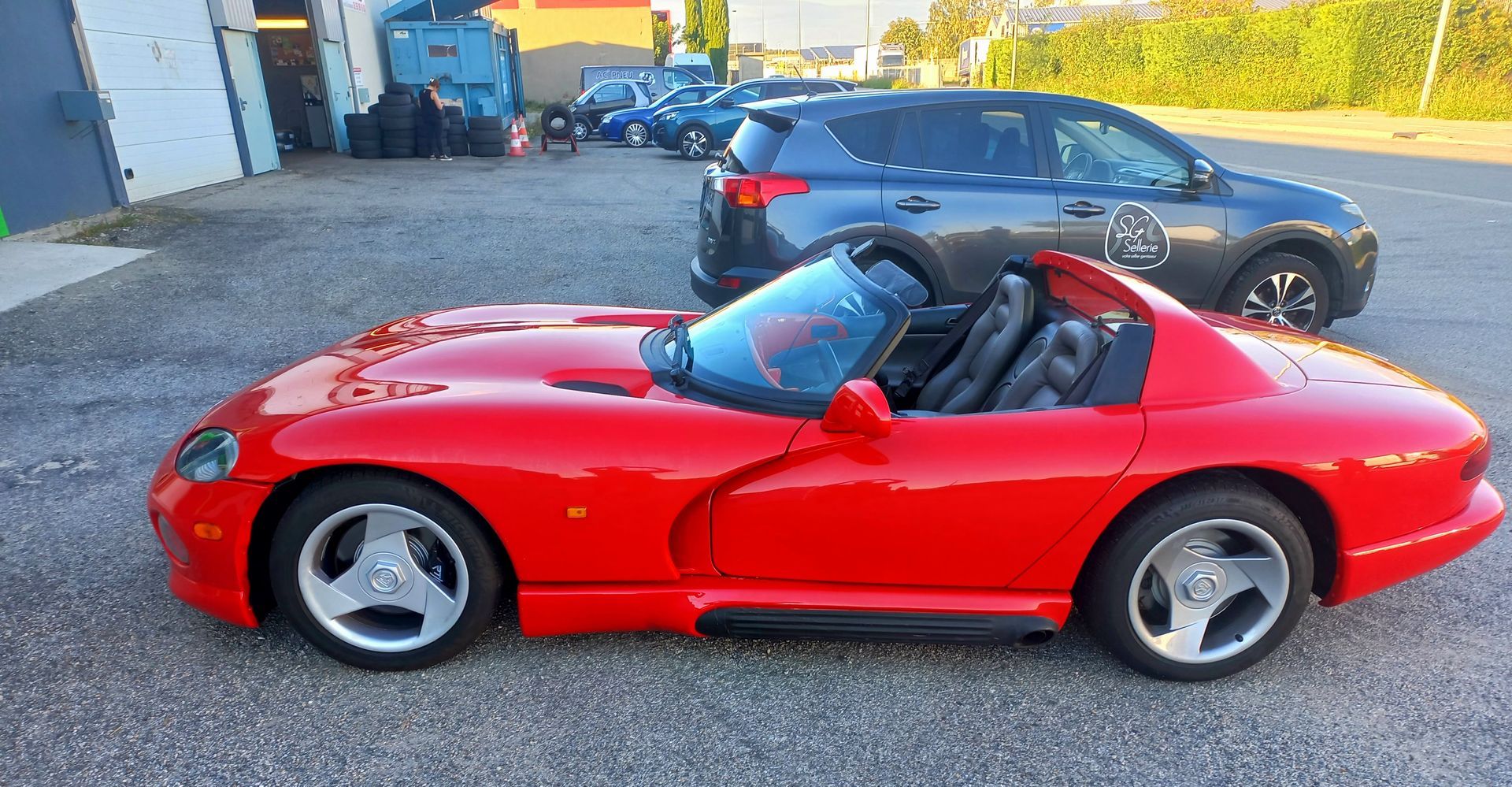 Voiture de sport décapotable rouge garée à l'extérieur, avec une berline noire en arrière-plan.