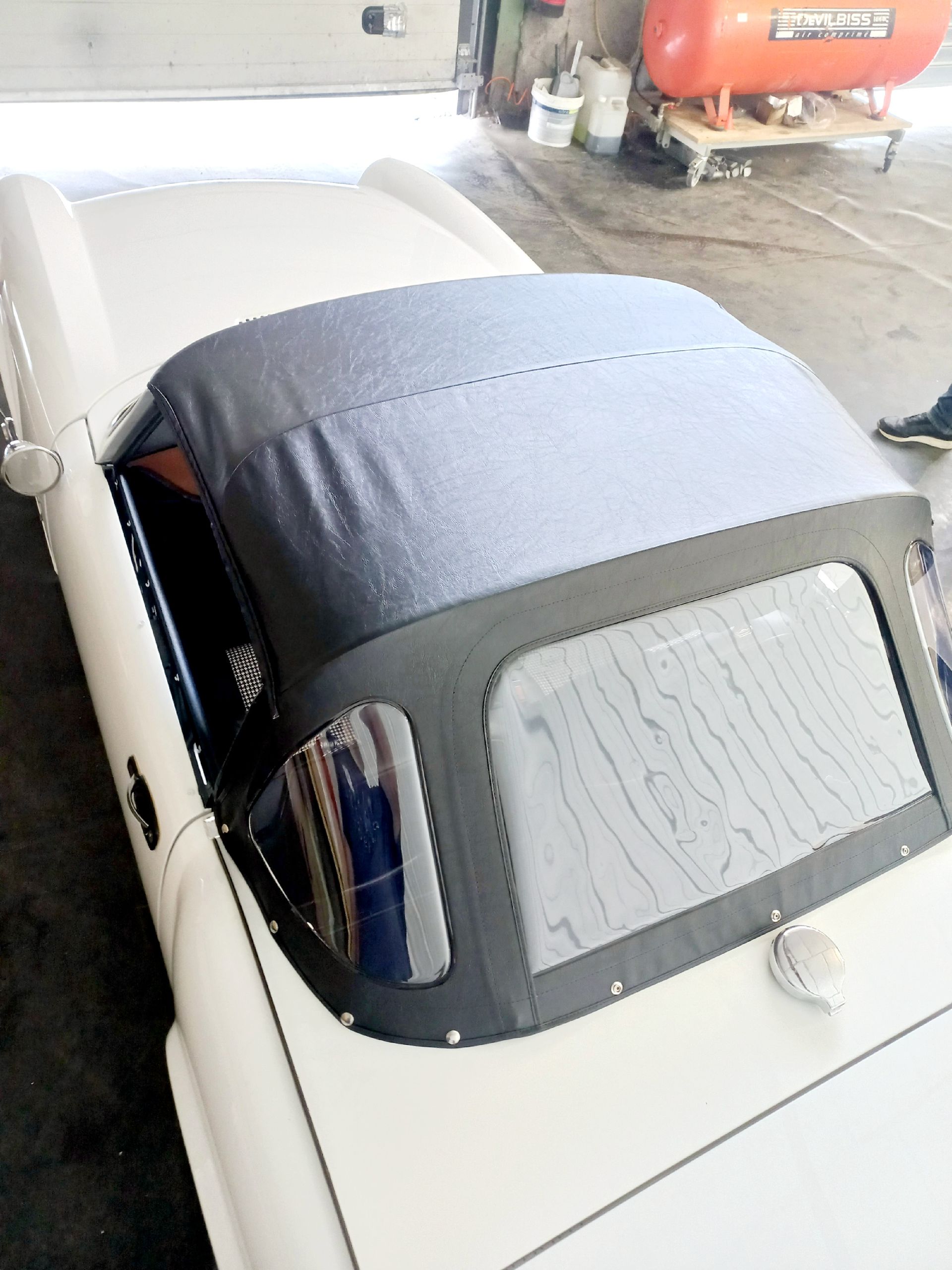 Un cabriolet vintage blanc avec une capote souple foncée est garé à l'intérieur, près du matériel d'atelier.