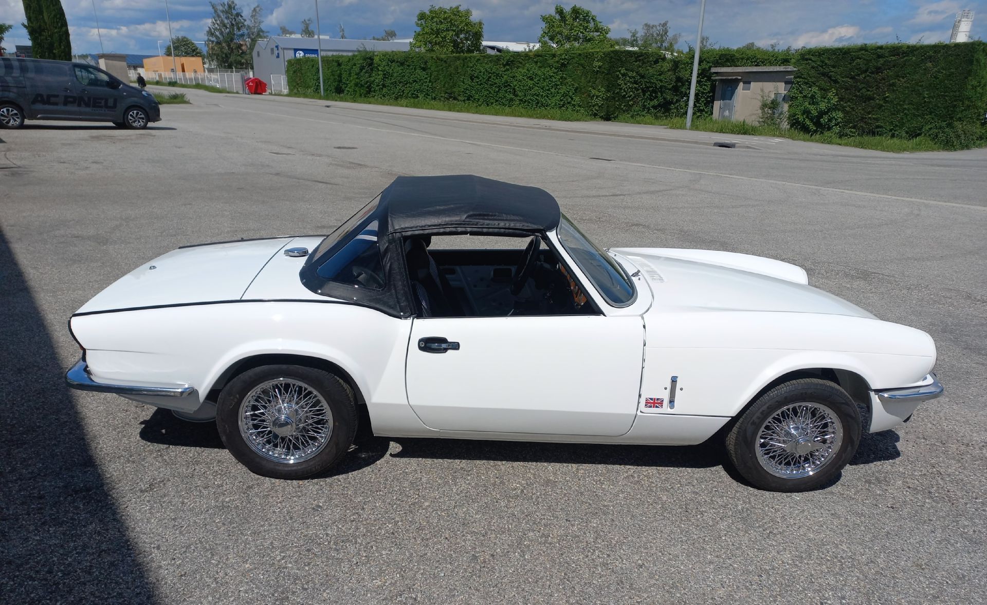 Voiture décapotable classique blanche avec capote noire garée sur un parking goudronné à l'extérieur.