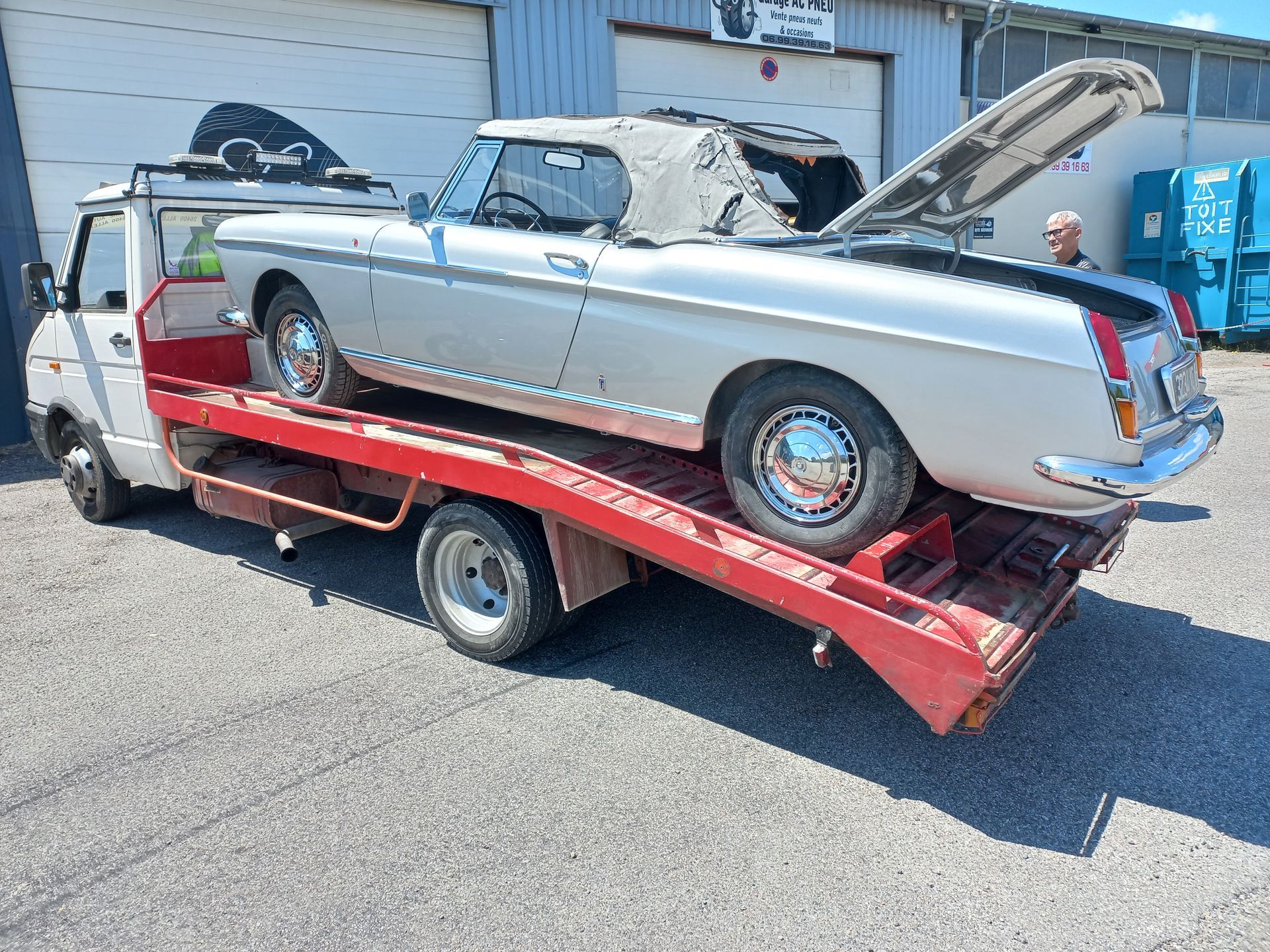 Voiture décapotable argentée sur une rampe de remorquage rouge devant un garage