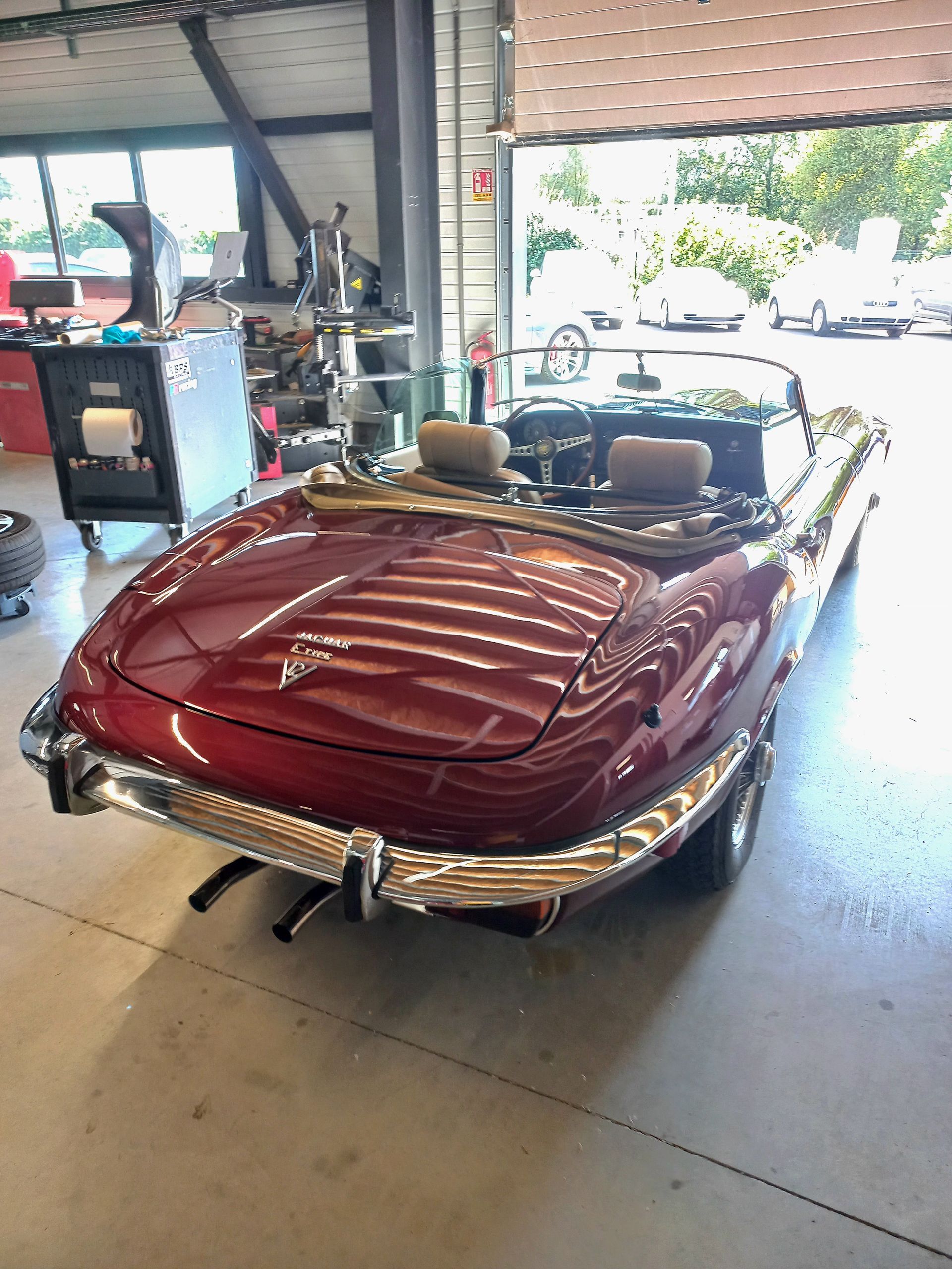 Cabriolet rouge vintage dans un garage, vue arrière avec pare-chocs chromé et sièges beiges, lumière du soleil filtrant par la portière ouverte