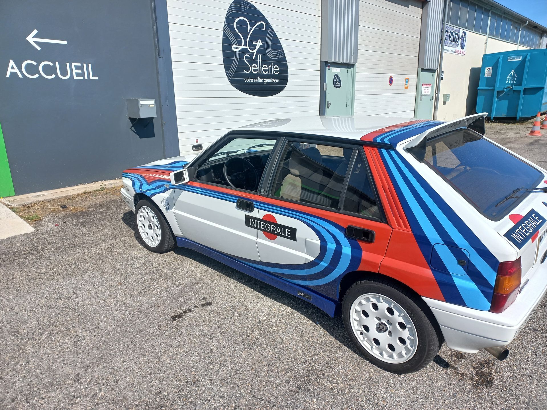 Une berline à hayon classique de style rallye avec des bandes de course rouges, bleues et blanches est garée devant un bâtiment portant l'inscription ACCUEIL.