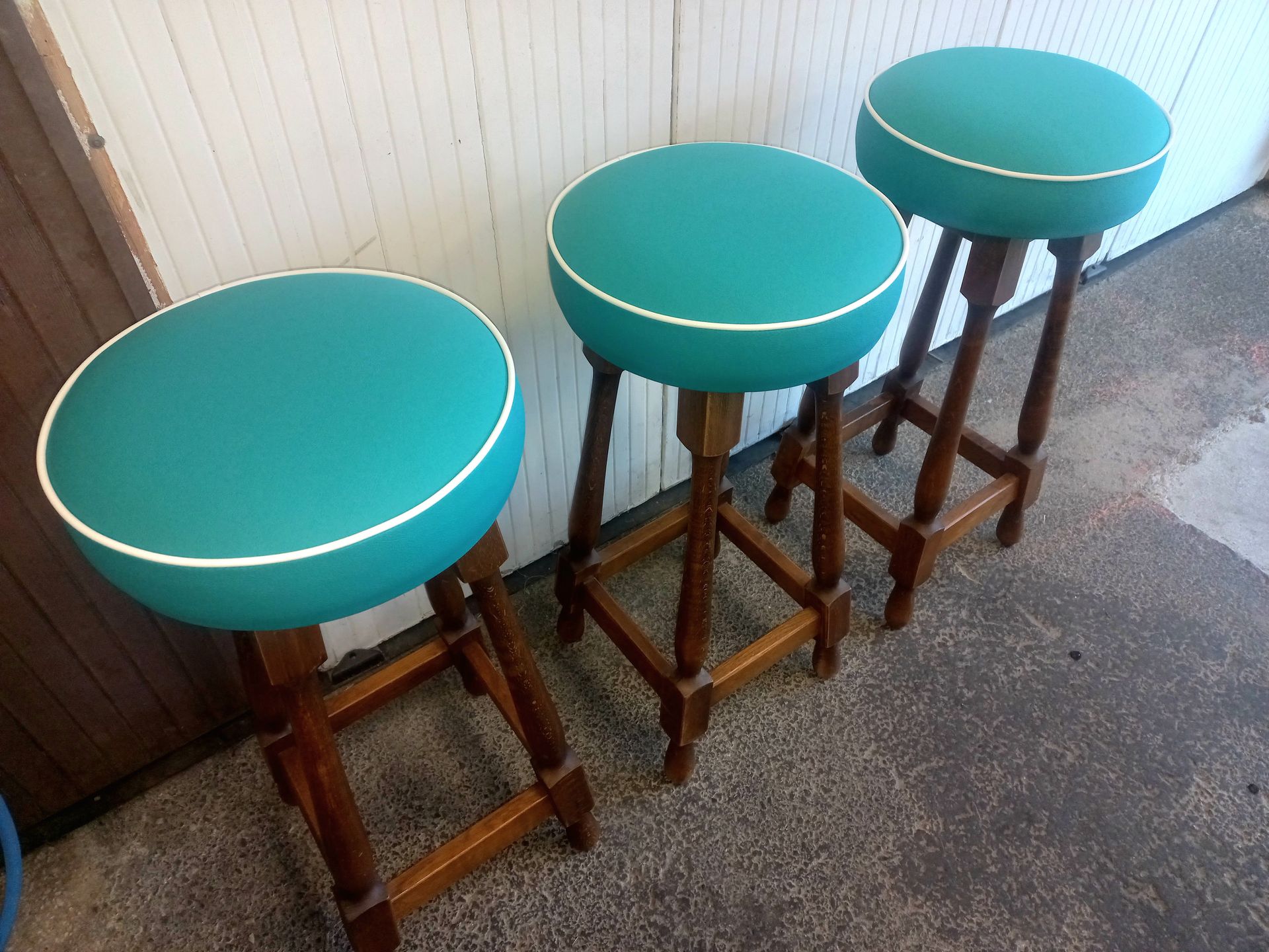 Trois tabourets bleu sarcelle à coussins et à structure en bois sont alignés contre un mur.