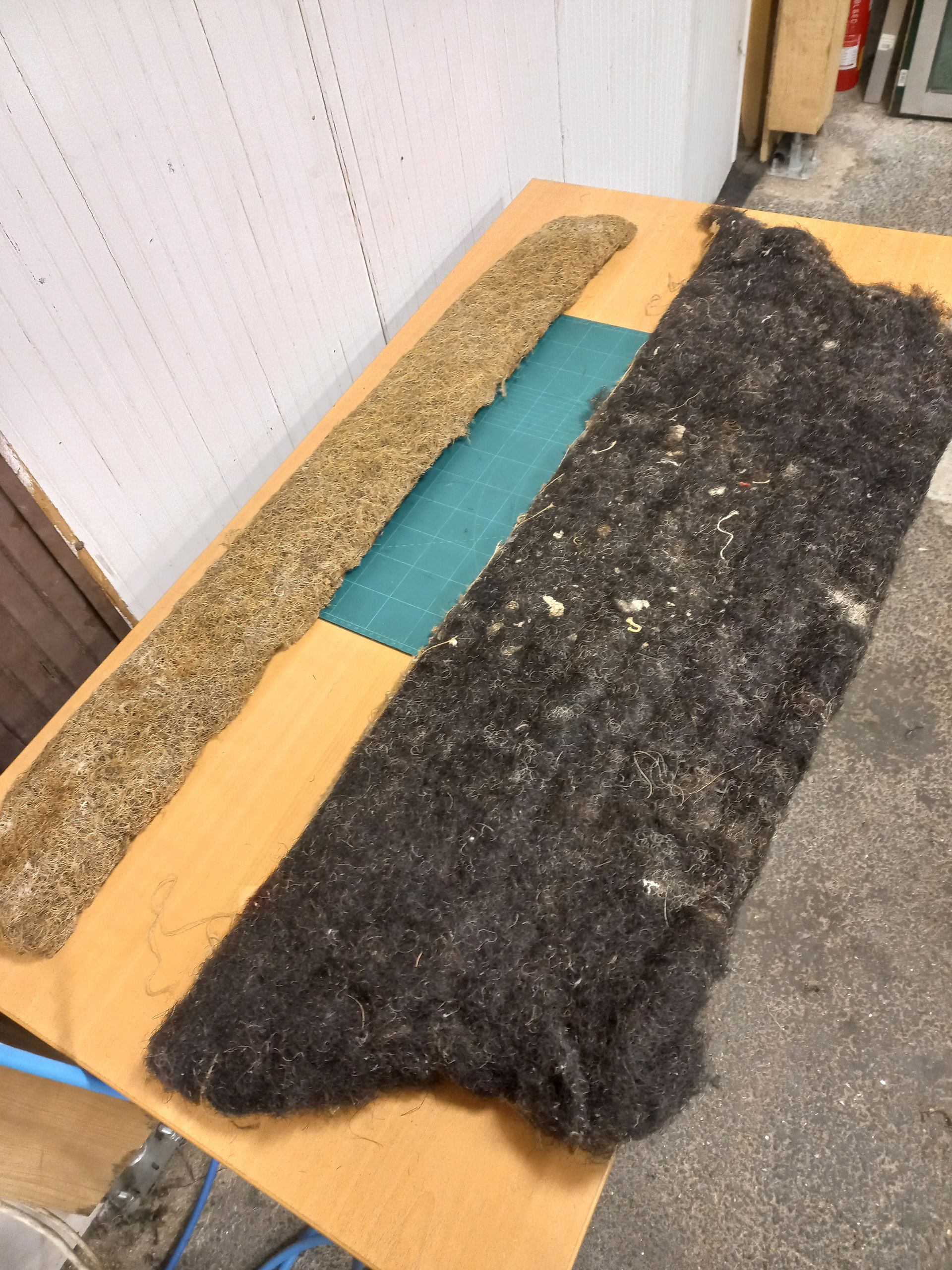 Deux longs tapis texturés, l'un beige et l'autre noir, posés sur une planche de bois à l'extérieur.