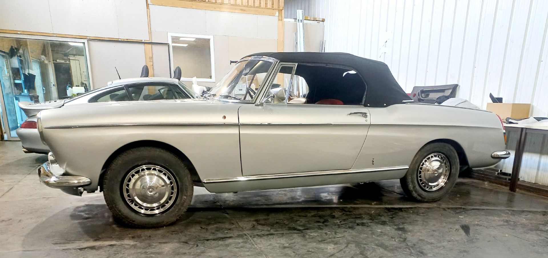 Voiture décapotable argentée classique garée dans un atelier, vue de côté avec capote noire.