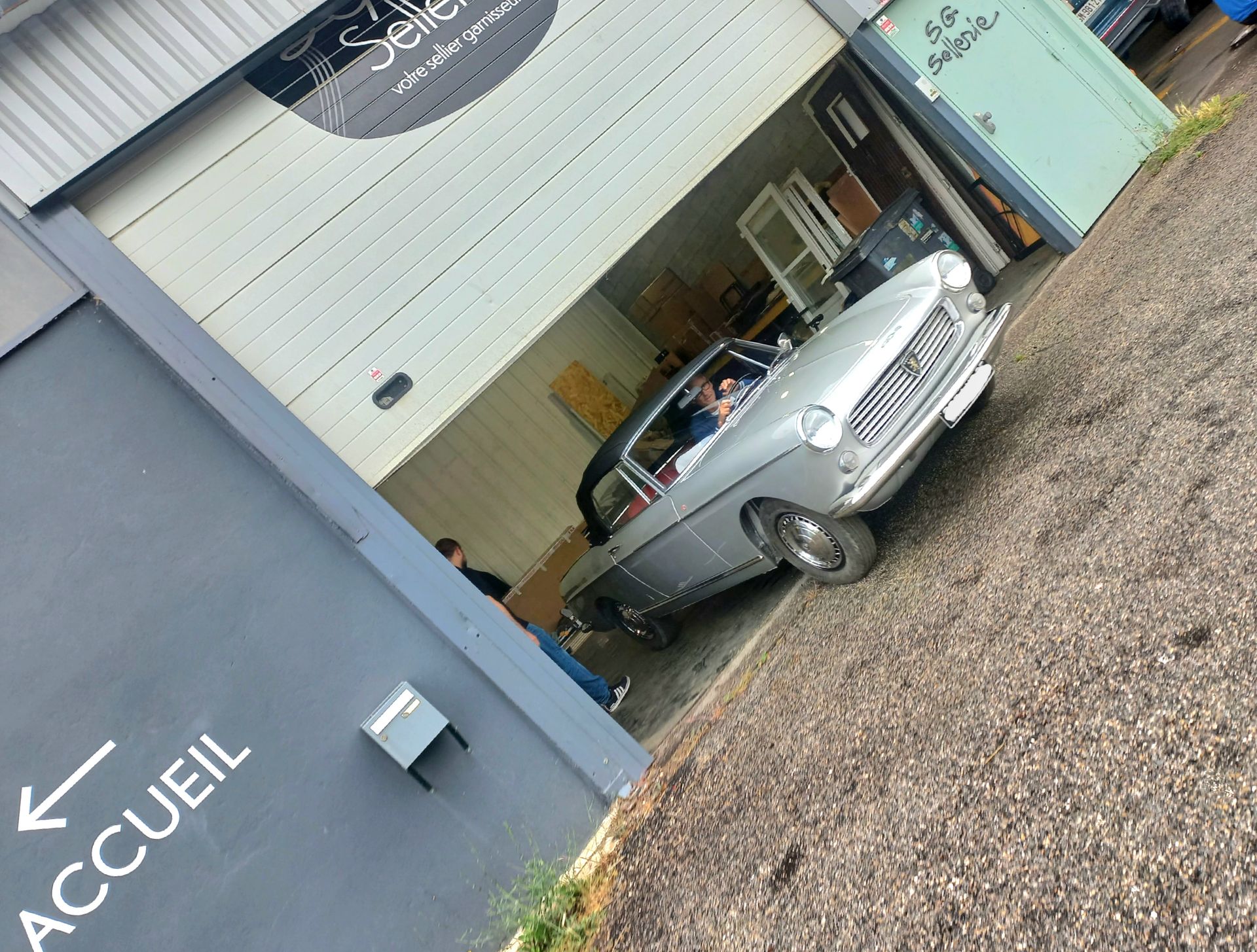 Garage ouvert avec une voiture classique argentée à l'intérieur, à côté d'un bâtiment bleu portant l'inscription Accueil.