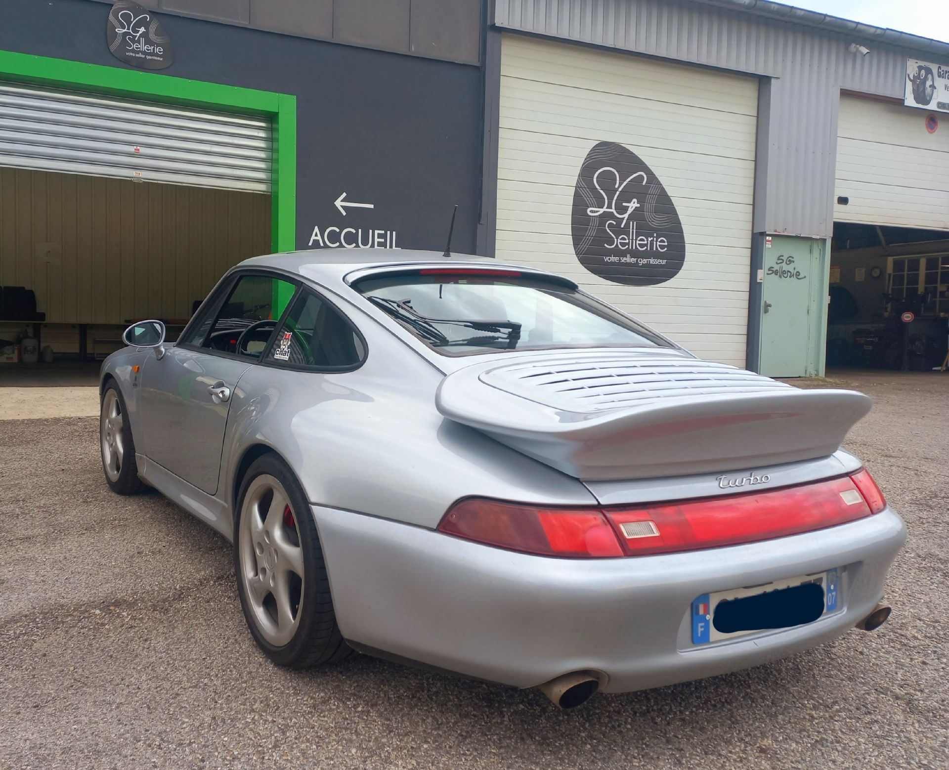 Porsche 911 argentée garée devant un garage, vue de trois quarts arrière, avec un grand aileron arrière.