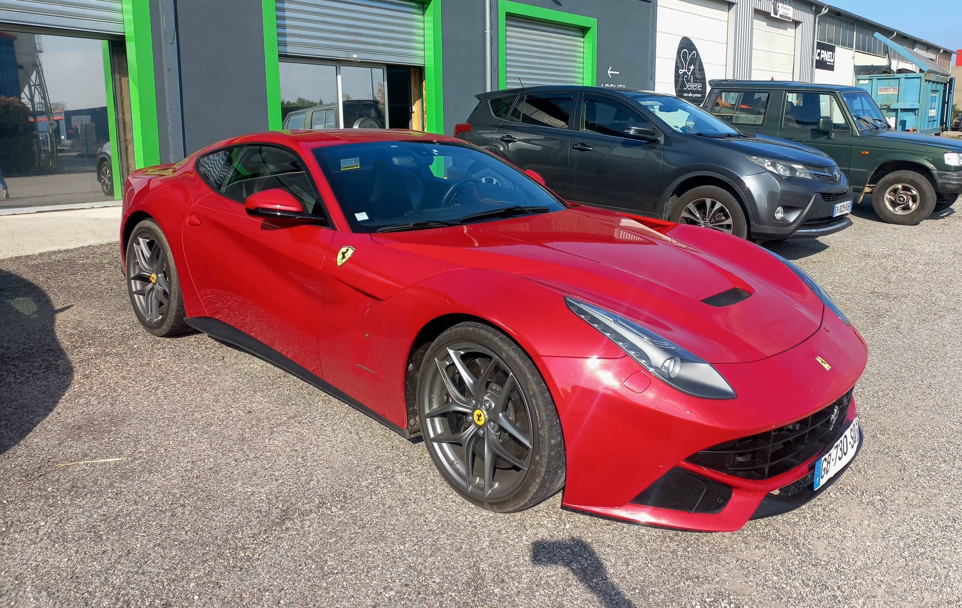 Une Ferrari rouge est garée devant un garage, avec un SUV sombre derrière.