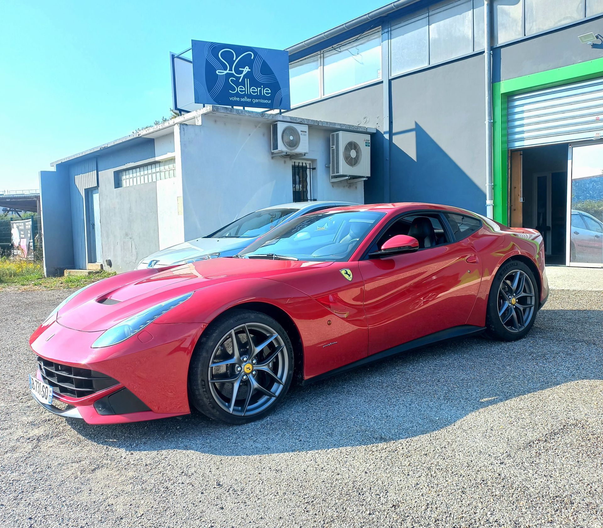 Voiture de sport rouge garée devant un garage, avec un bâtiment gris en arrière-plan.