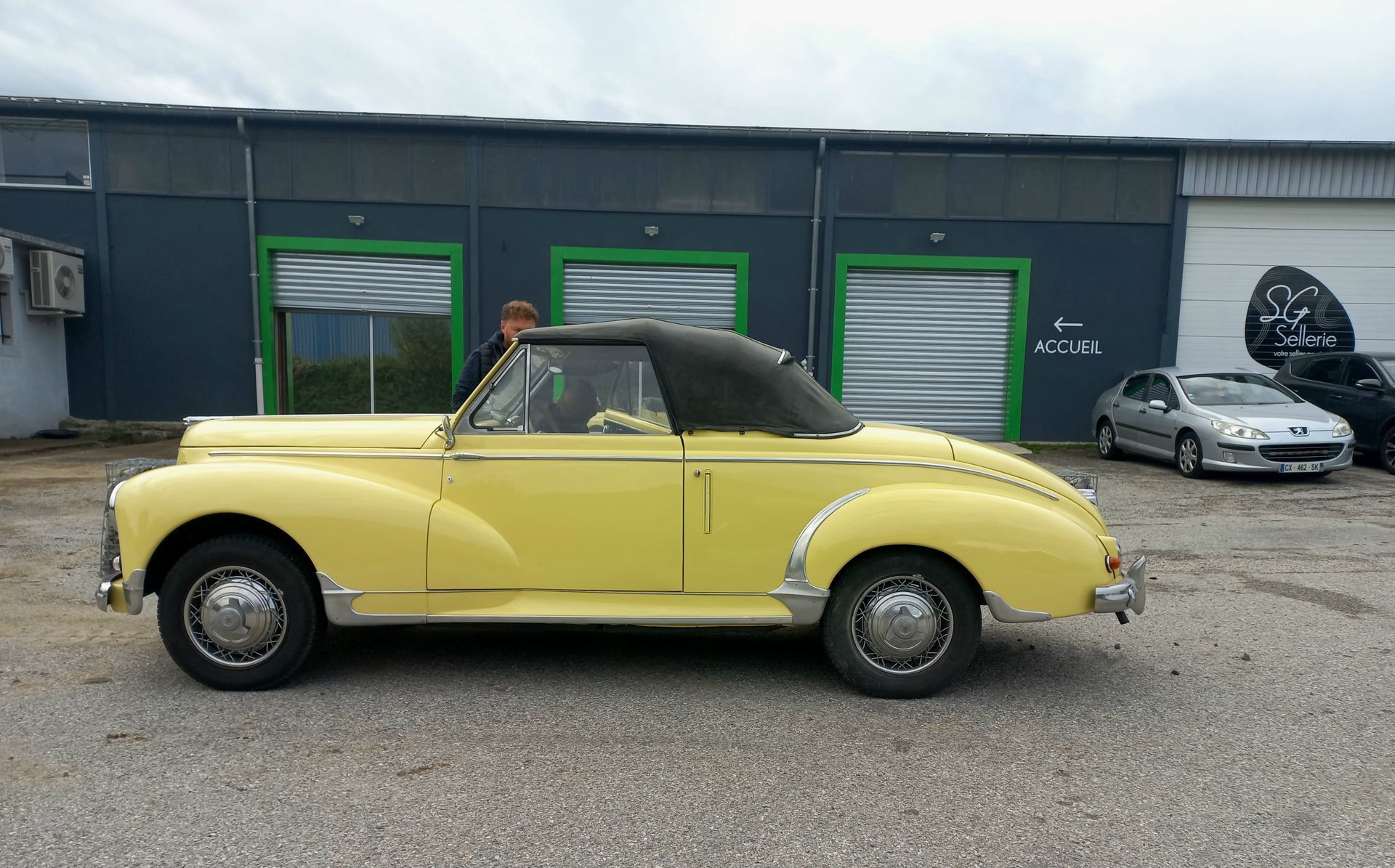 Une décapotable jaune vintage garée devant un garage, avec une voiture de sport argentée en arrière-plan.
