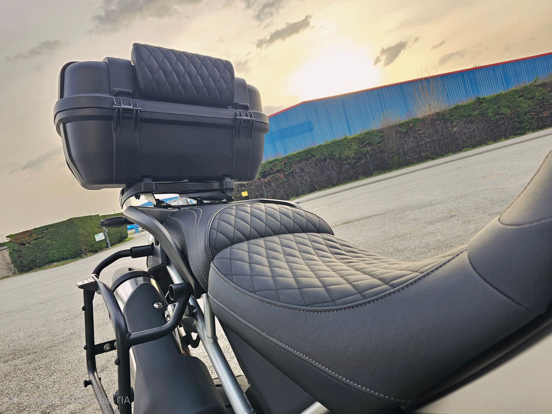 Moto noire avec selle matelassée et coffre arrière, garée sur une route au coucher du soleil