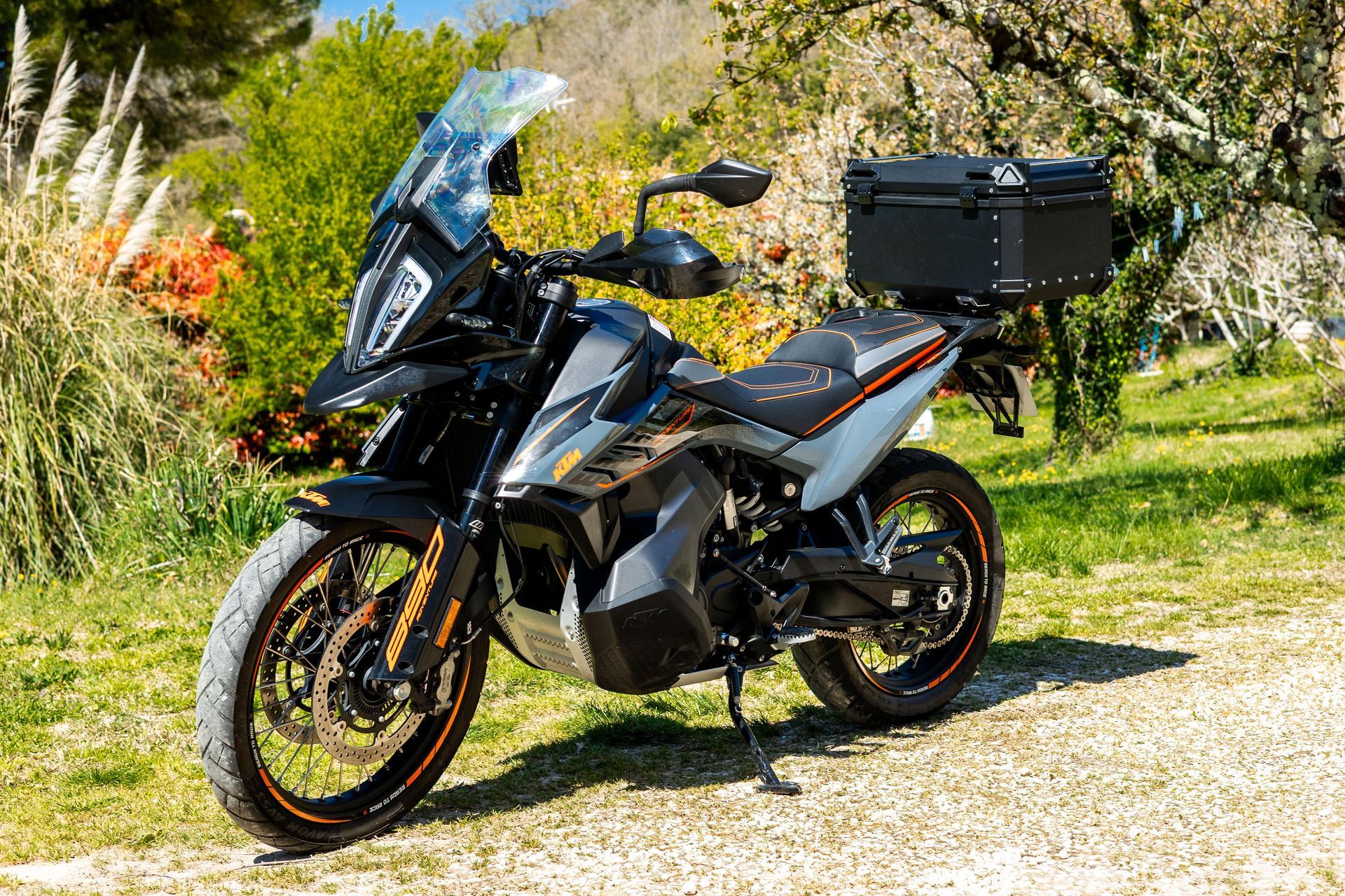 Une moto de tourisme noire est garée sur du gravier dans un jardin ensoleillé, arboré et fleuri.