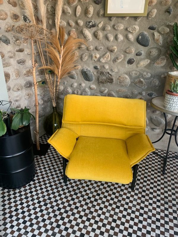 Fauteuil jaune à assise rabattable dans une pièce rustique avec mur de galets et sol carrelé noir et blanc