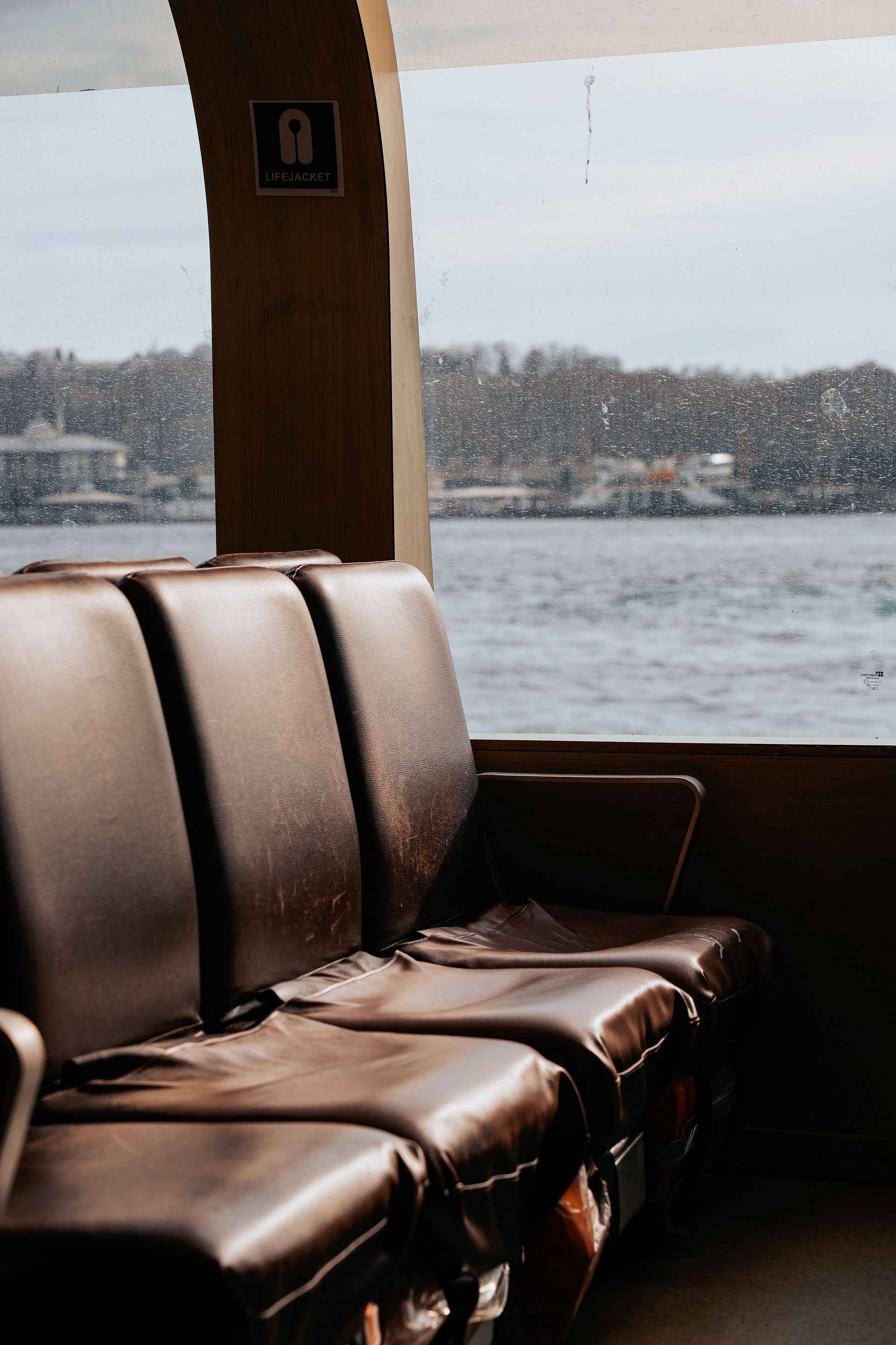 Sièges en cuir marron à l'intérieur d'un ferry, avec un rivage enneigé visible par la fenêtre.