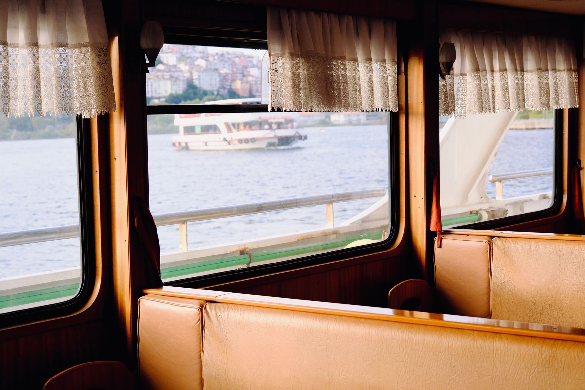 Intérieur d'un ferry avec des sièges beiges et des fenêtres à rideaux donnant sur l'eau et un autre bateau