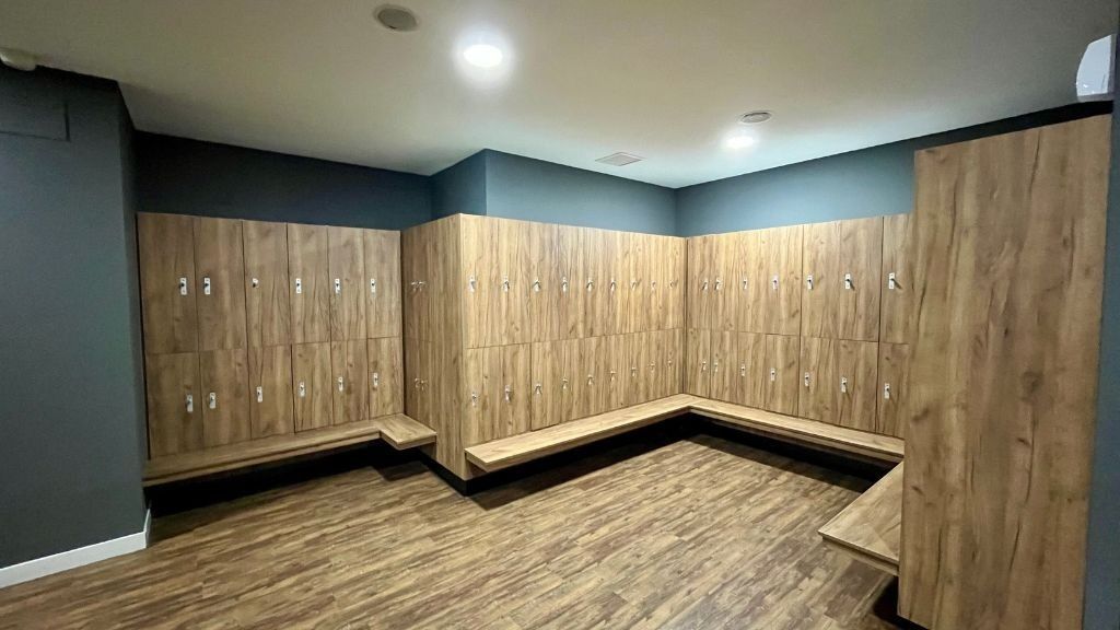 Vestuario con taquillas y bancos de madera contra una pared gris y suelo marrón.