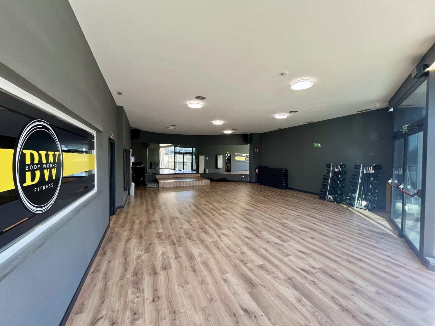 Estudio de baile vacío con piso de apariencia de madera, logotipo grande, puertas de vidrio e iluminación empotrada.