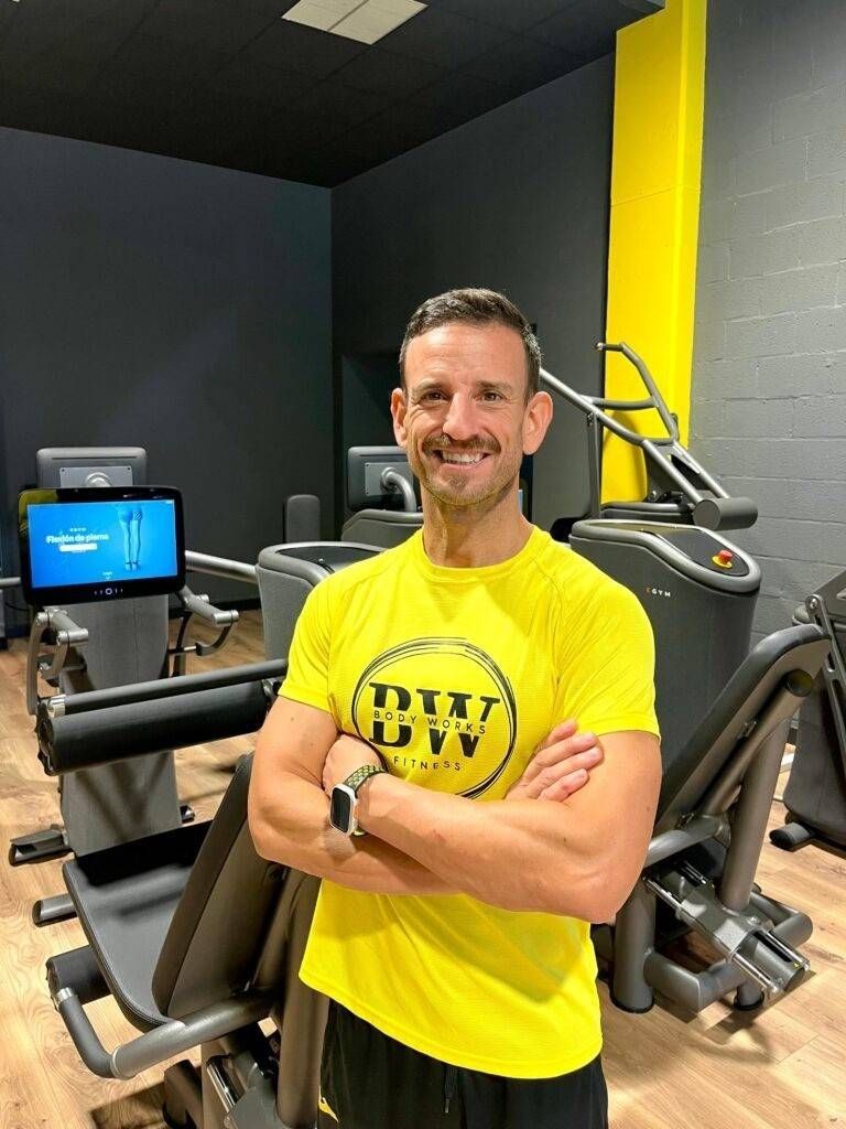 Hombre con camisa amarilla, con los brazos cruzados, sonríe en un gimnasio, rodeado de máquinas de ejercicios.