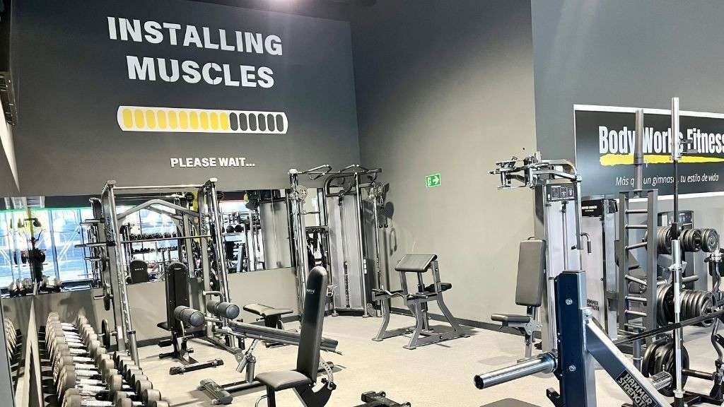 Interior de gimnasio con varias máquinas de pesas y un gráfico de "Instalando músculos" en la pared.