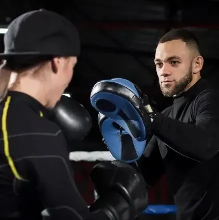 Boxeador practicando golpes con un entrenador sosteniendo guantes en un gimnasio, ropa negra.