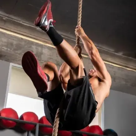 Hombre trepando una cuerda en un gimnasio, con las piernas envueltas alrededor de la cuerda, zapatillas rojas.