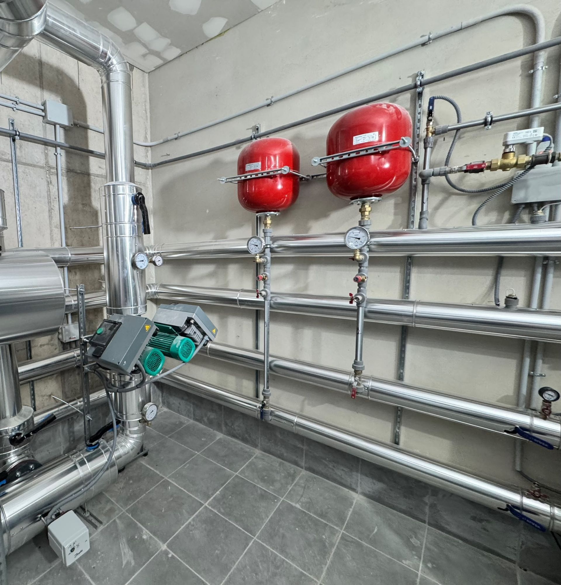 Sala de equipos industriales con tuberías metálicas, dos depósitos de presión rojos y suelo de baldosas