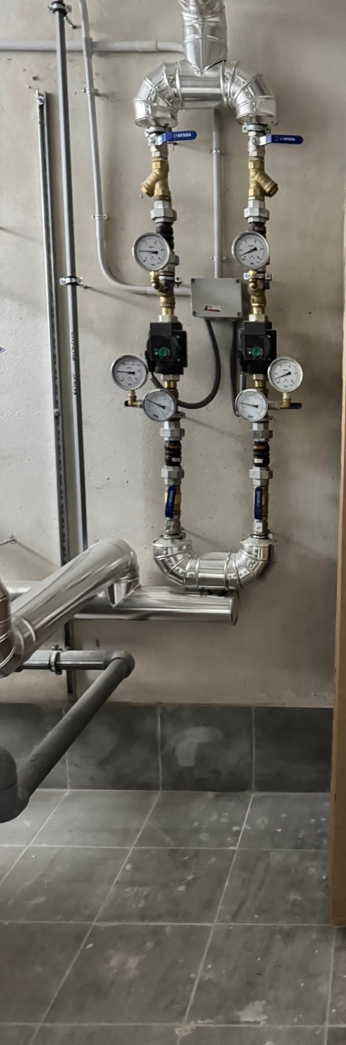 Tuberías metálicas, válvulas y medidores instalados en una pared de hormigón en una sala de instalaciones industriales.