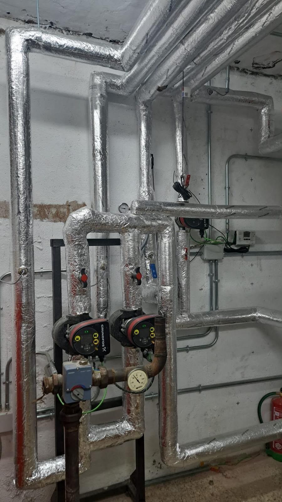 Tuberías y dos bombas industriales montadas en la pared de una sala de servicios, con conductos metálicos y manómetros.