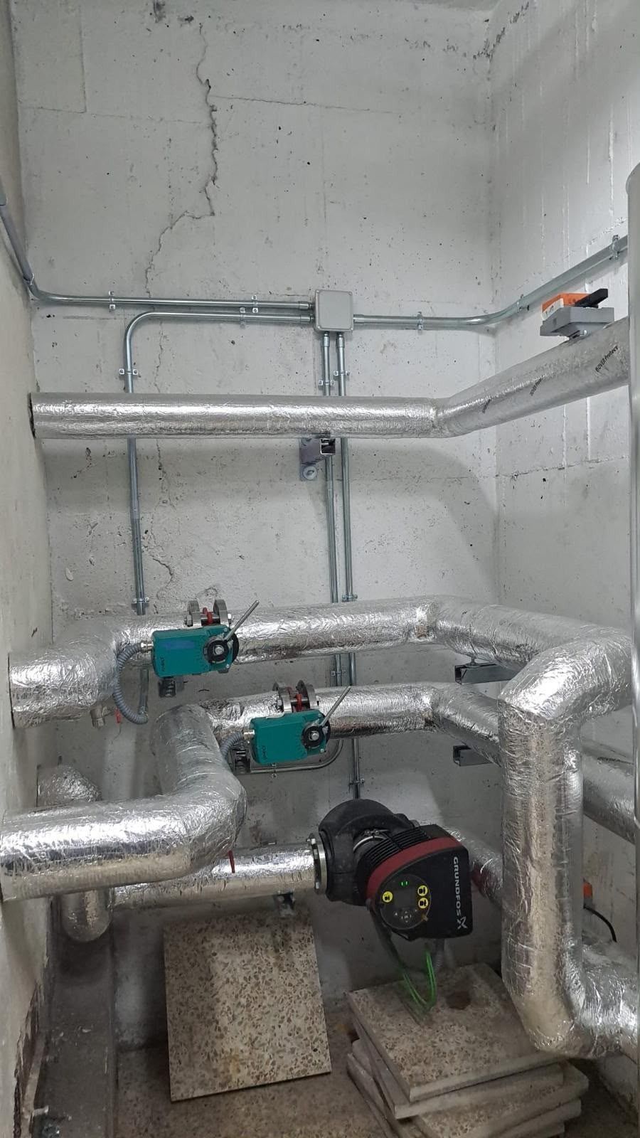 Tuberías industriales aisladas y válvulas en una sala de instalaciones con paredes de hormigón.