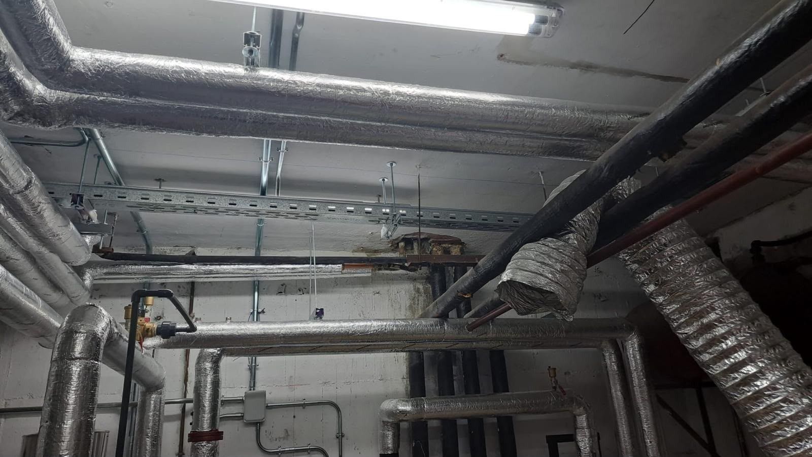 Techo industrial con conductos plateados a la vista, tuberías y luces fluorescentes en un cuarto de servicio