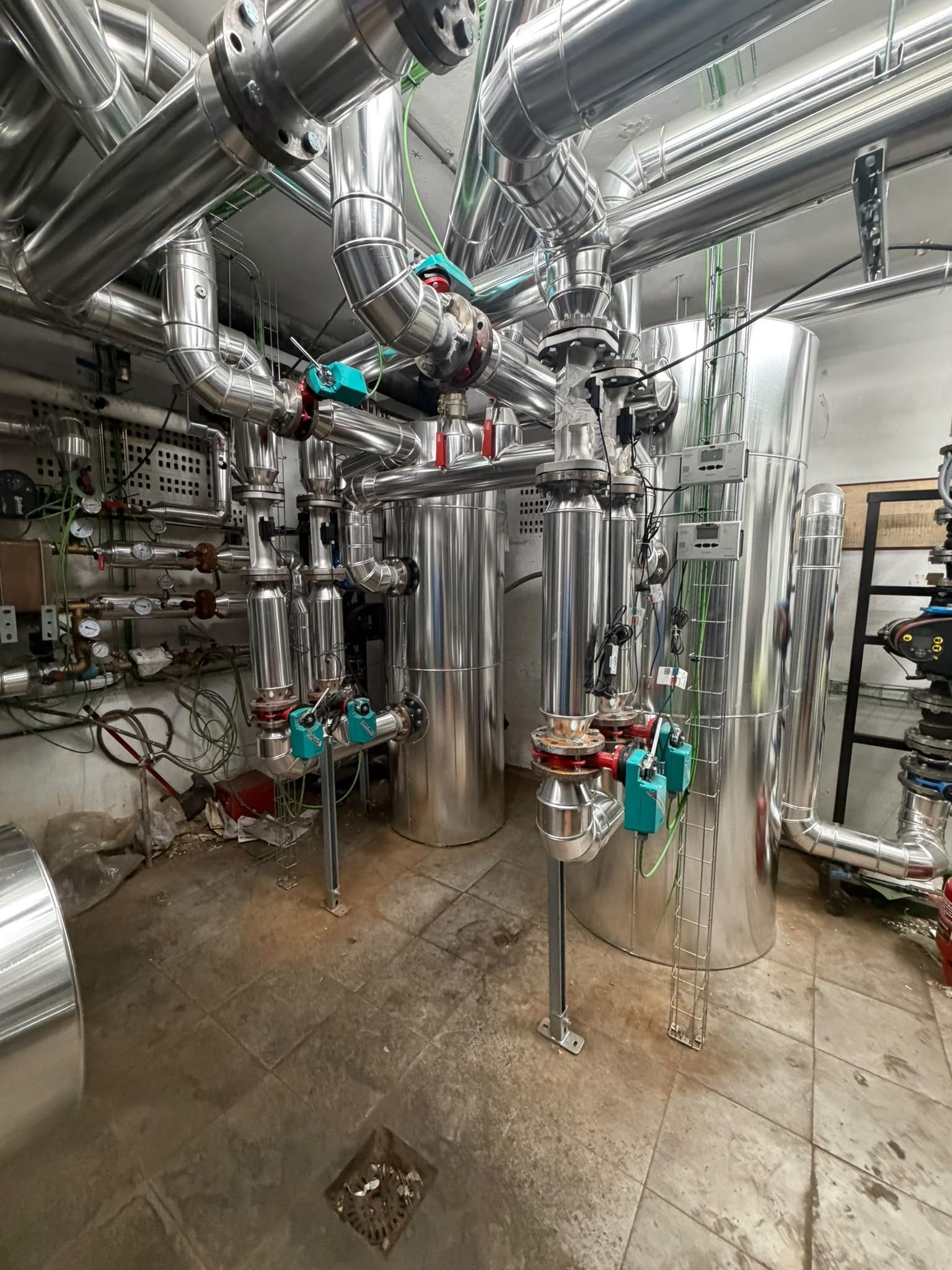 Sala de instalaciones industriales con tanques, tuberías y válvulas de acero inoxidable sobre un suelo de hormigón mojado