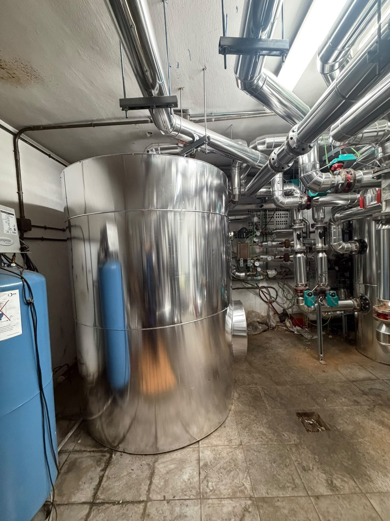 Sótano industrial con un gran depósito de acero inoxidable, un cilindro azul y tuberías y maquinaria a la vista.