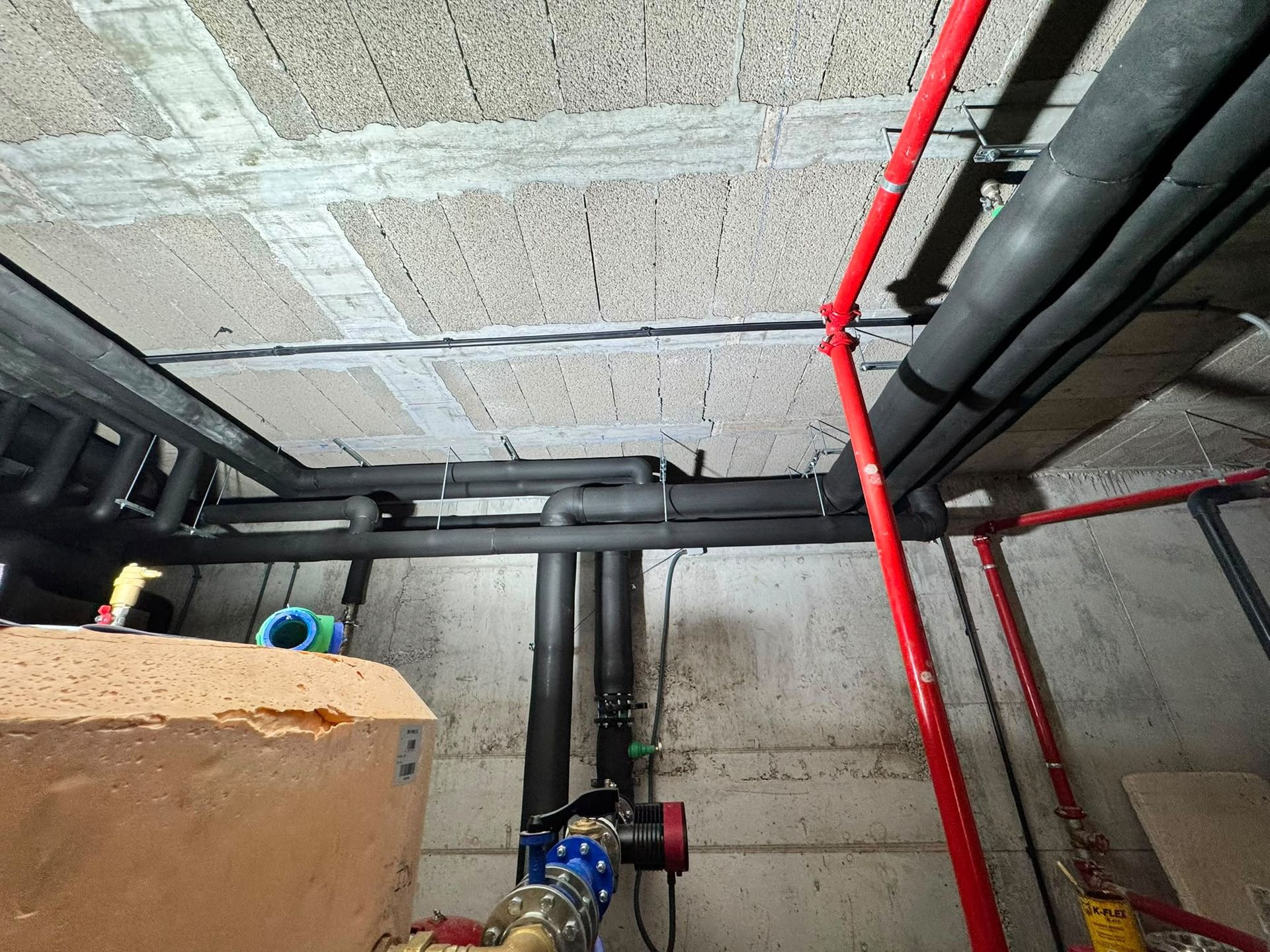 Techo del cuarto de instalaciones del sótano, con tuberías a la vista, un conducto rojo y un medidor de agua