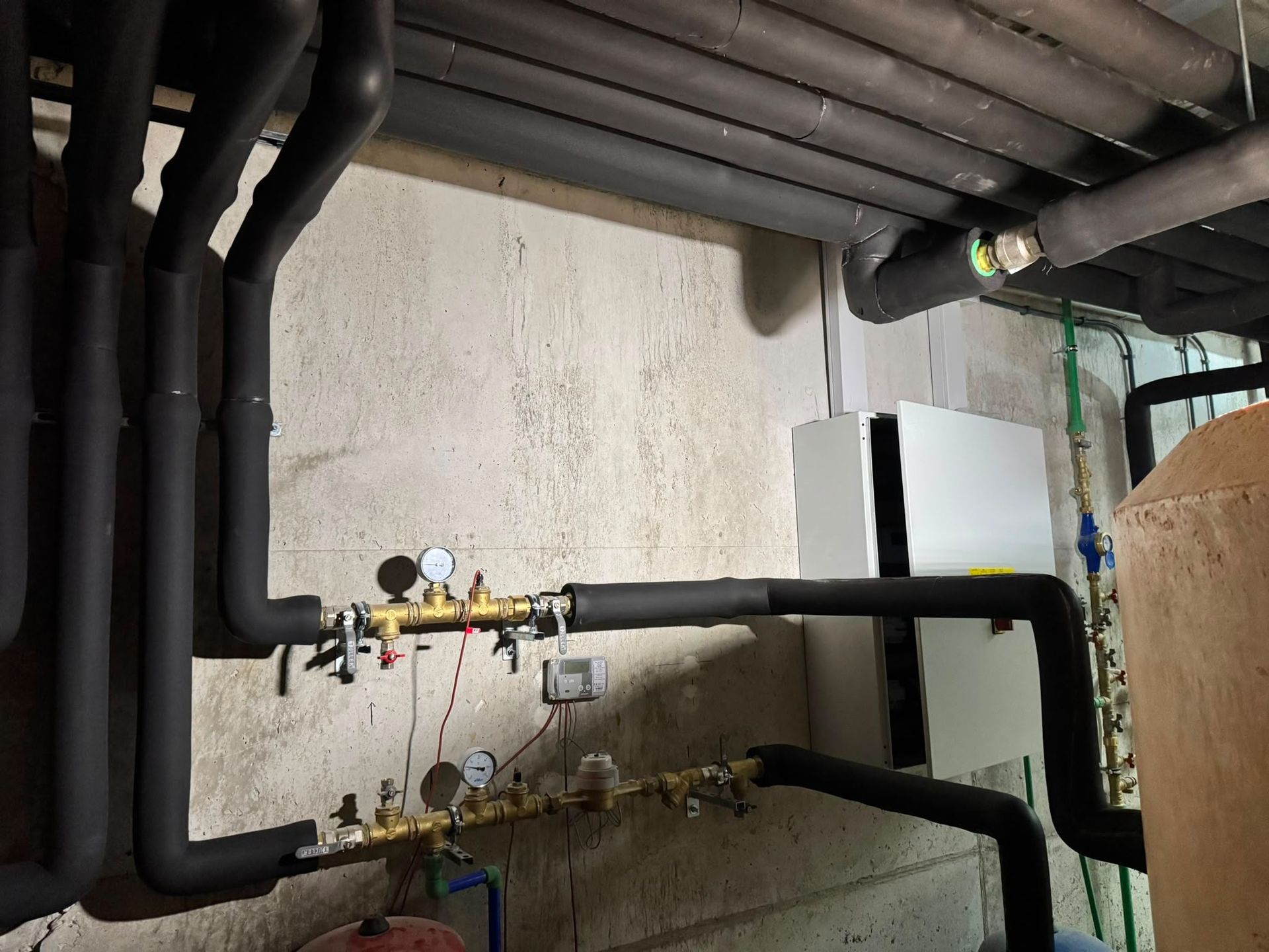 Tuberías y válvulas de servicios en una pared del sótano, con tuberías negras aisladas y manómetros.