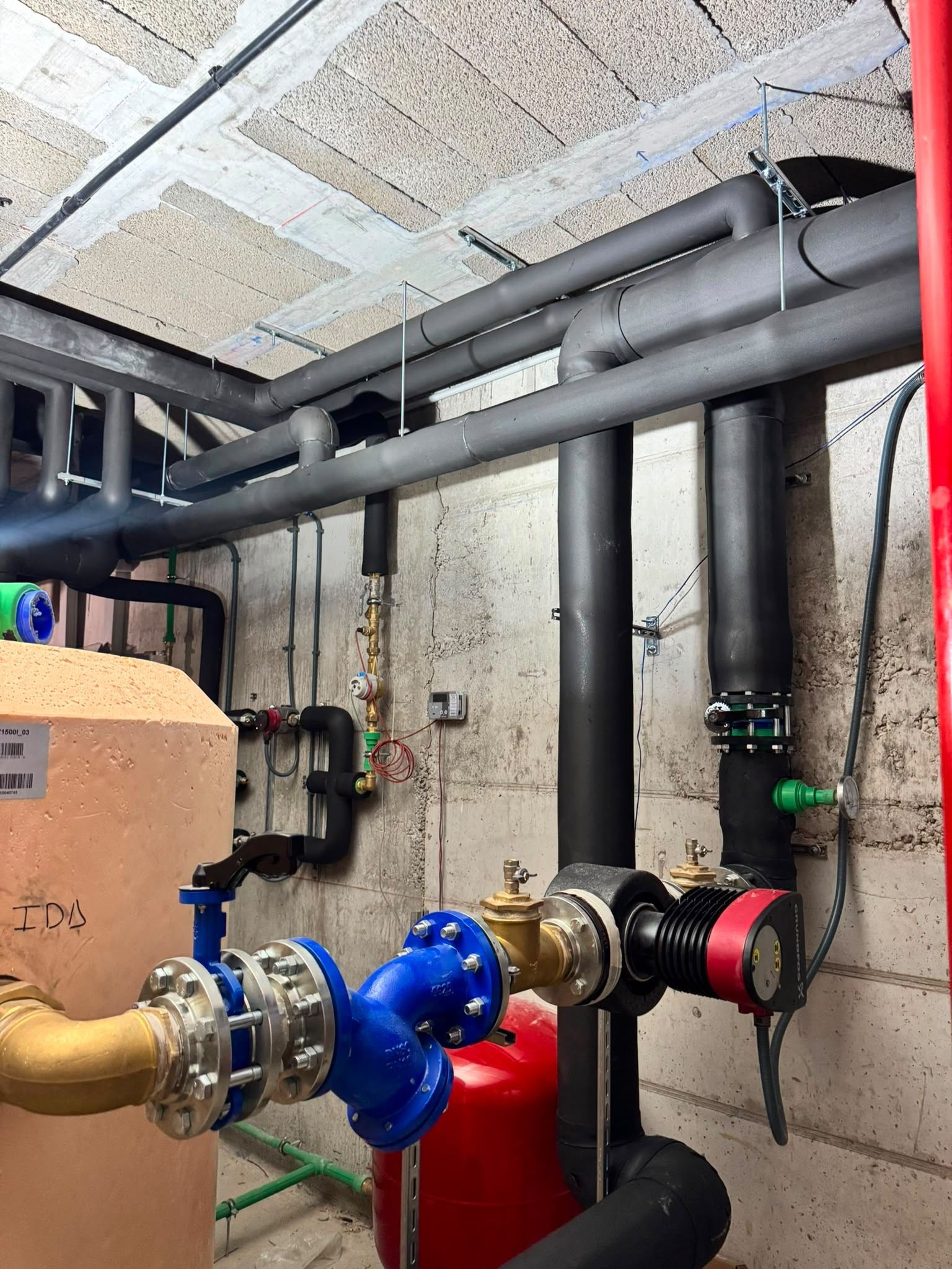 Tuberías industriales, válvulas y manómetros en una sala de servicios con techo y paredes de hormigón.