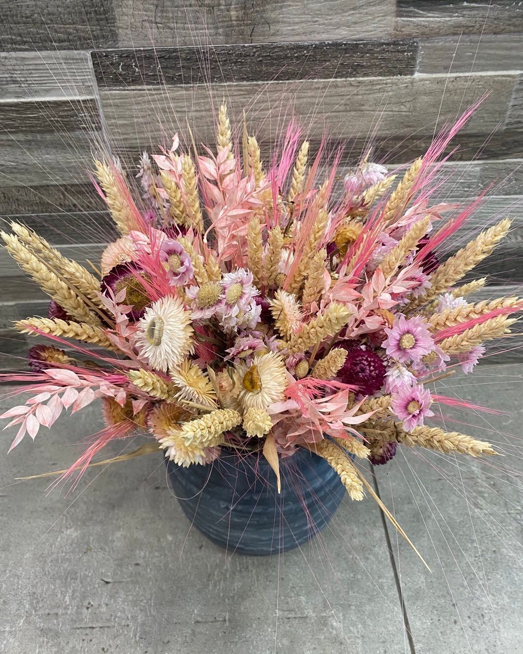 Composition de fleurs séchées dans un vase texturé bleu foncé. Des épis de blé beige et des fleurs roses sont agencés avec finesse.