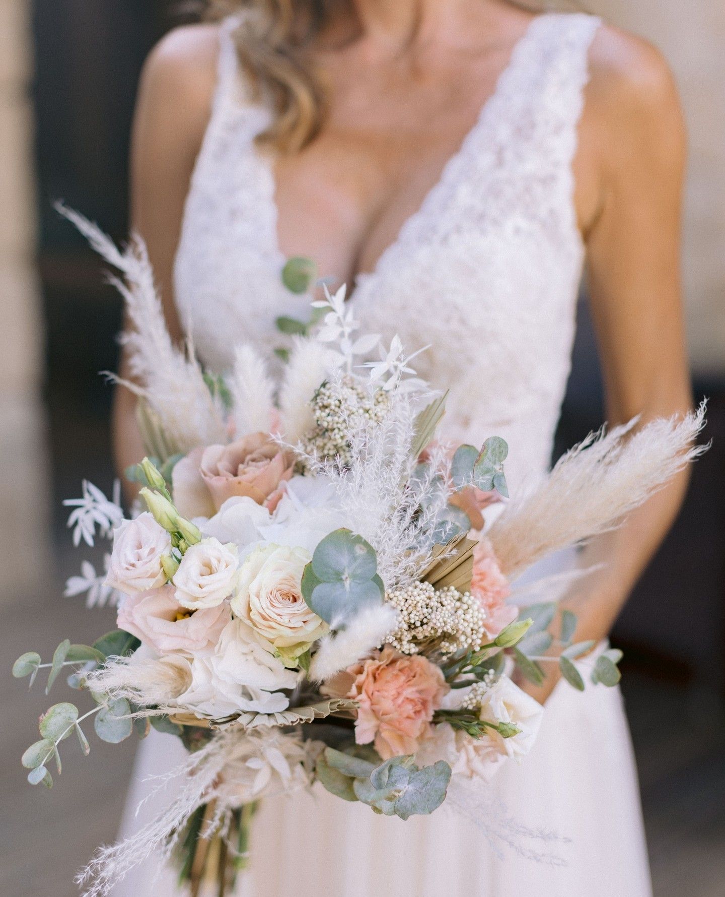 La mariée tient un bouquet de fleurs pêche, blanches et vertes ; elle porte une robe en dentelle blanche.