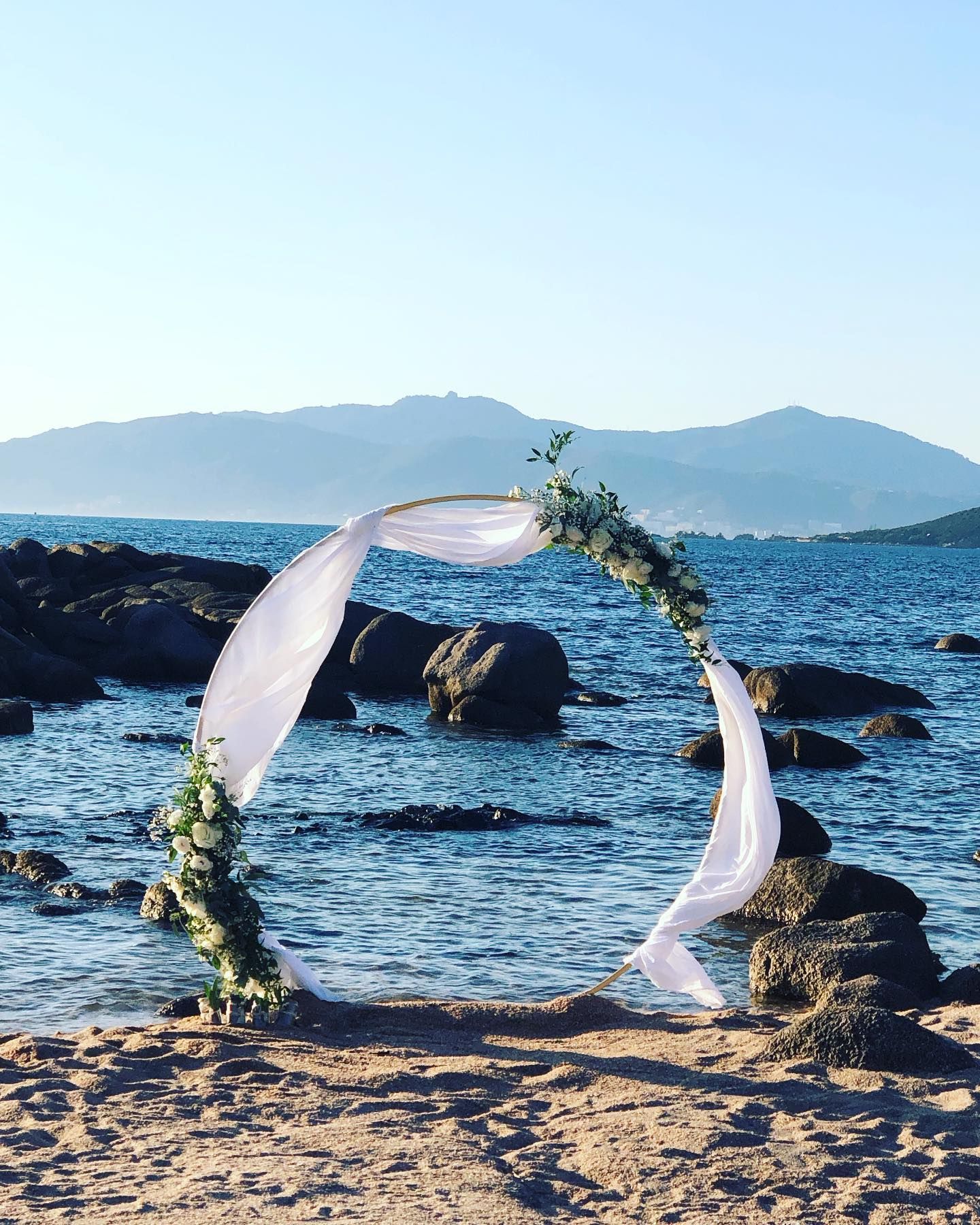 Arche de mariage circulaire sur une plage de sable, décorée de tissus et de verdure, avec vue sur la mer et les montagnes.