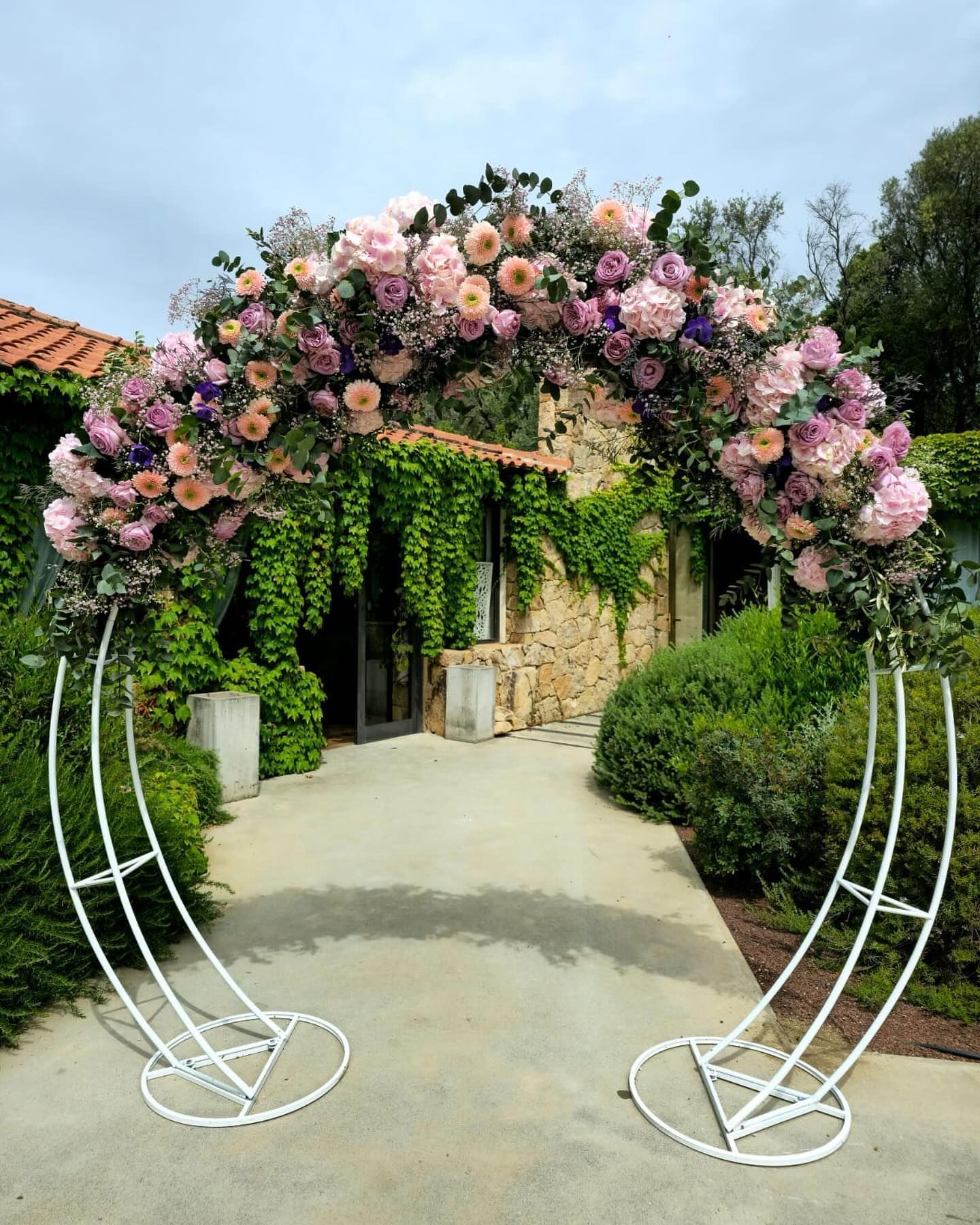 Arche florale blanche ornée de fleurs roses, pêche et violettes, située dans un jardin avec un bâtiment en pierre.