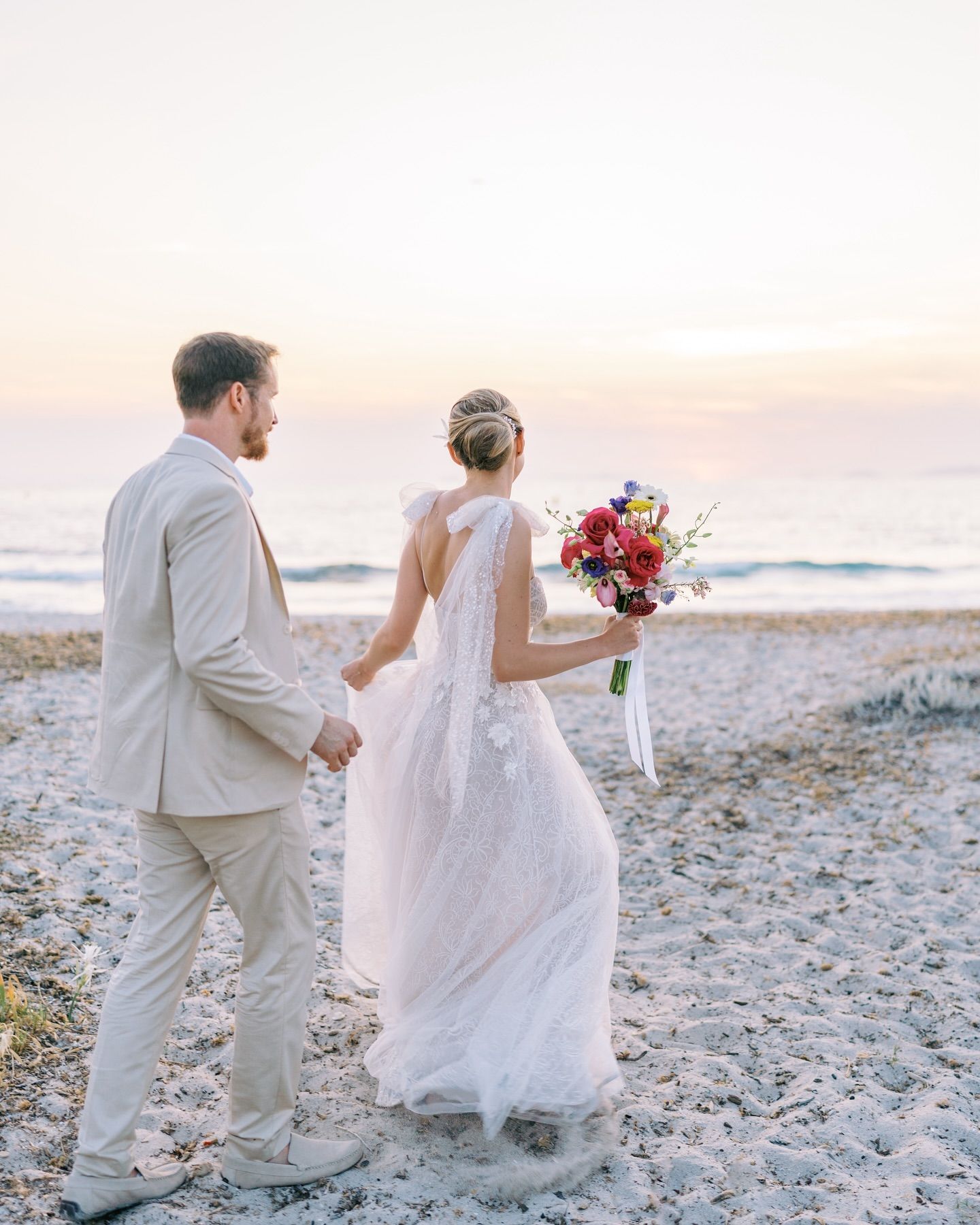 Les mariés se promènent main dans la main sur une plage au coucher du soleil. La mariée tient un bouquet coloré.