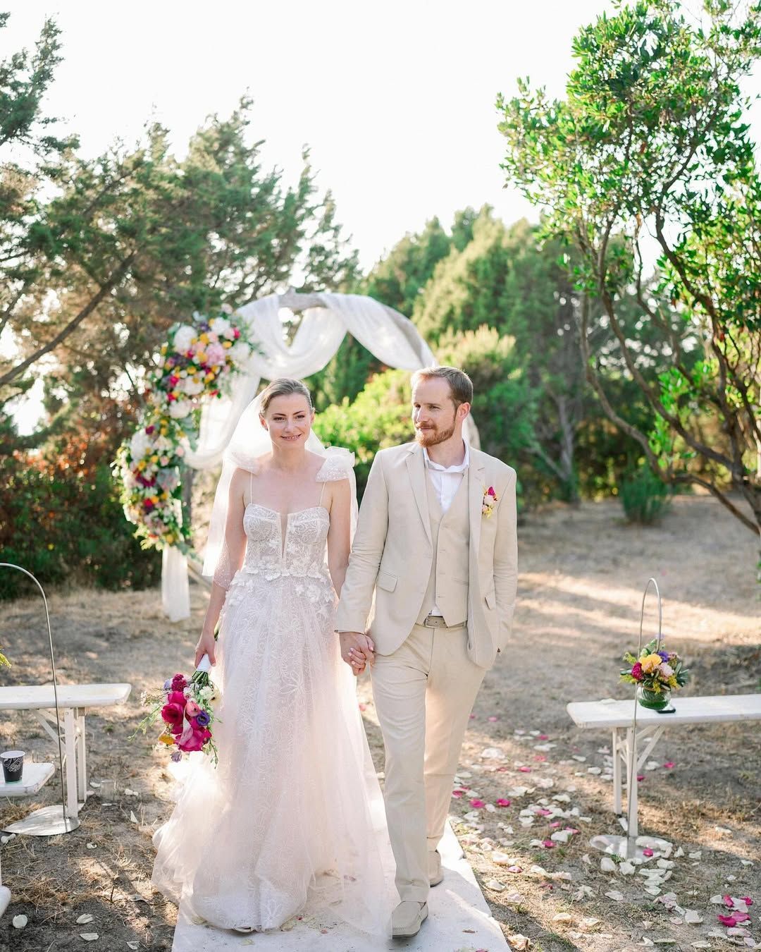 Les mariés marchent main dans la main dans l'allée centrale, en direction de la caméra, après une cérémonie en extérieur.