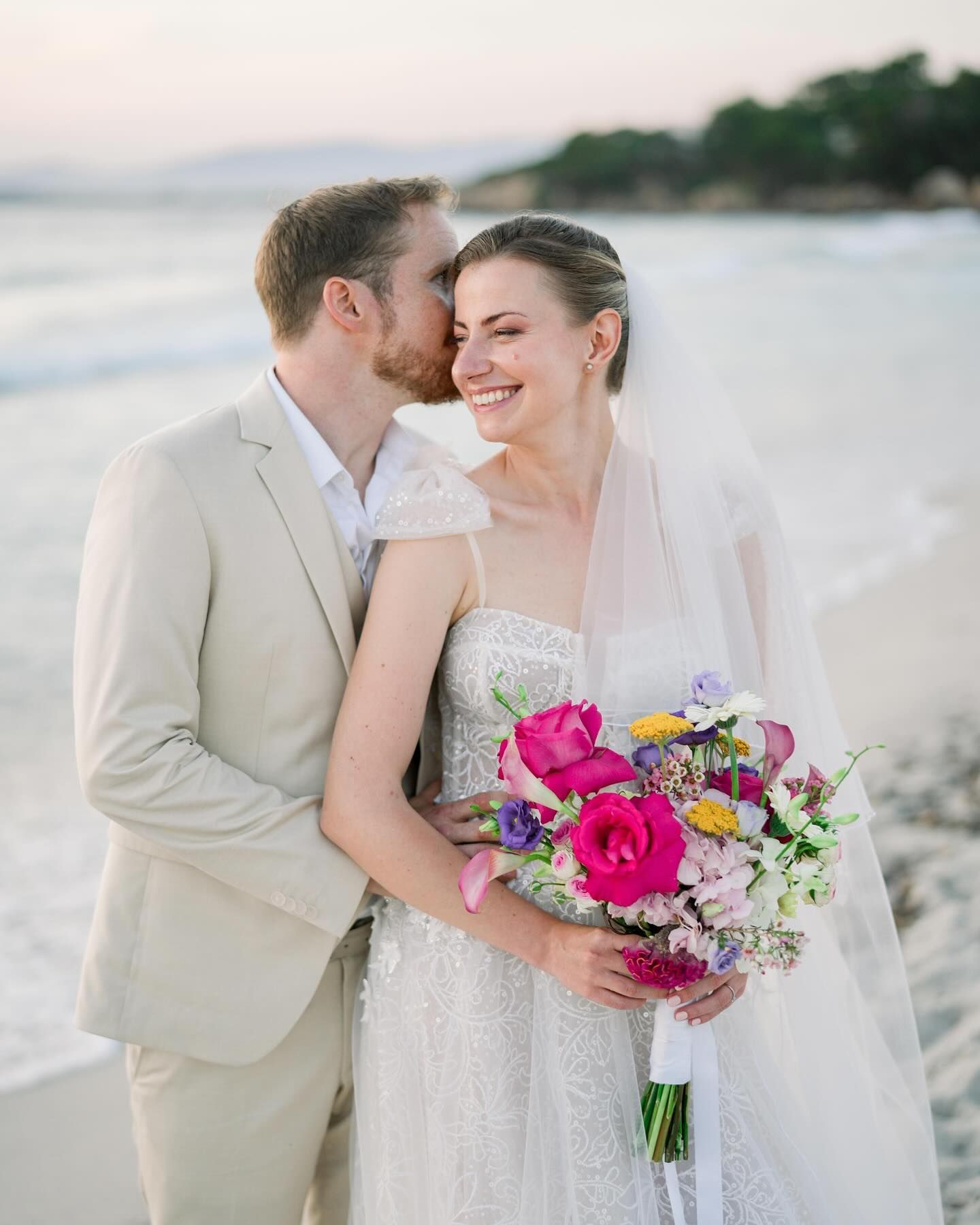 Un couple sur la plage au coucher du soleil ; la mariée en robe tient son bouquet, le marié l'embrasse sur la joue.