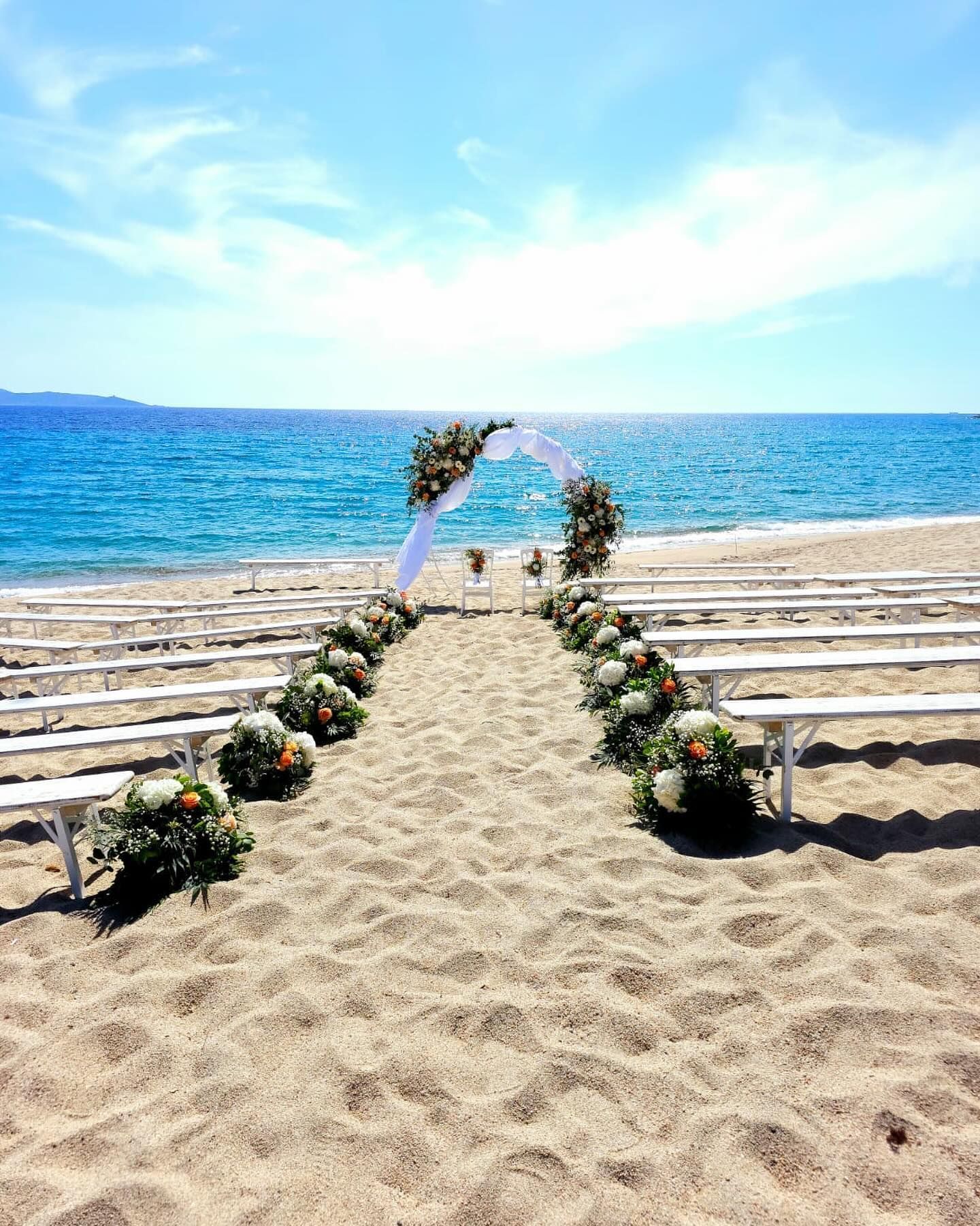 Cérémonie de mariage sur la plage, avec bancs blancs, arche fleurie et l'océan en arrière-plan.