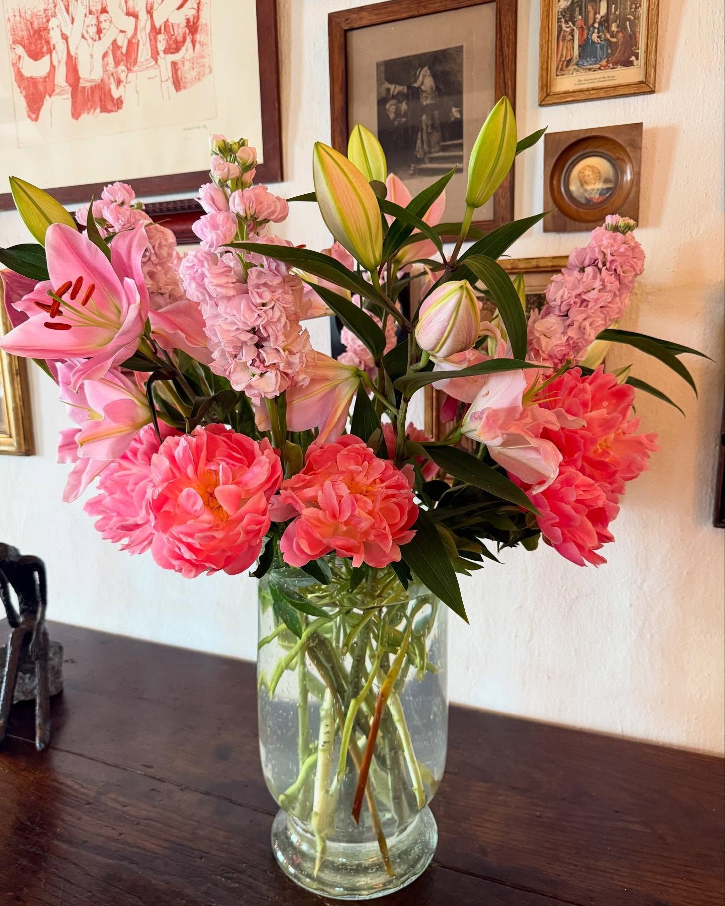 Un bouquet éclatant de lys roses, de pivoines et d'autres fleurs dans un vase en verre posé sur une surface en bois sombre.