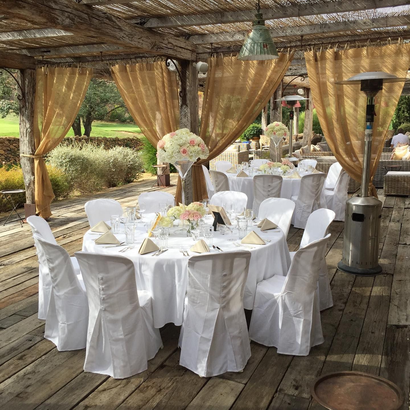Tables de réception en extérieur, nappes blanches, chaises, centres de table floraux, rideaux dorés, terrasse en bois, pergola.