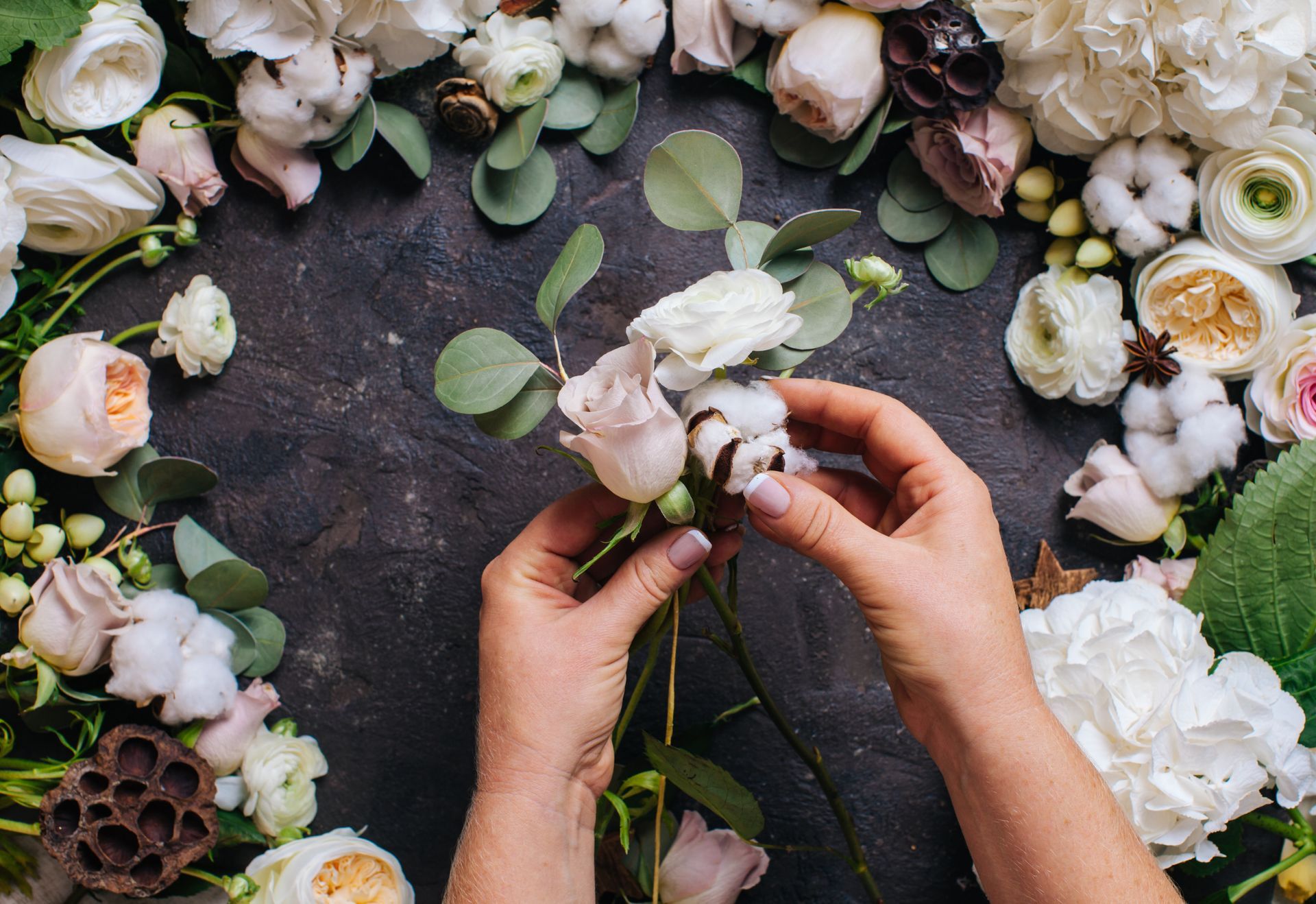 Des mains arrangent un bouquet de fleurs rose pâle et blanches sur une surface sombre, entourées de fleurs.
